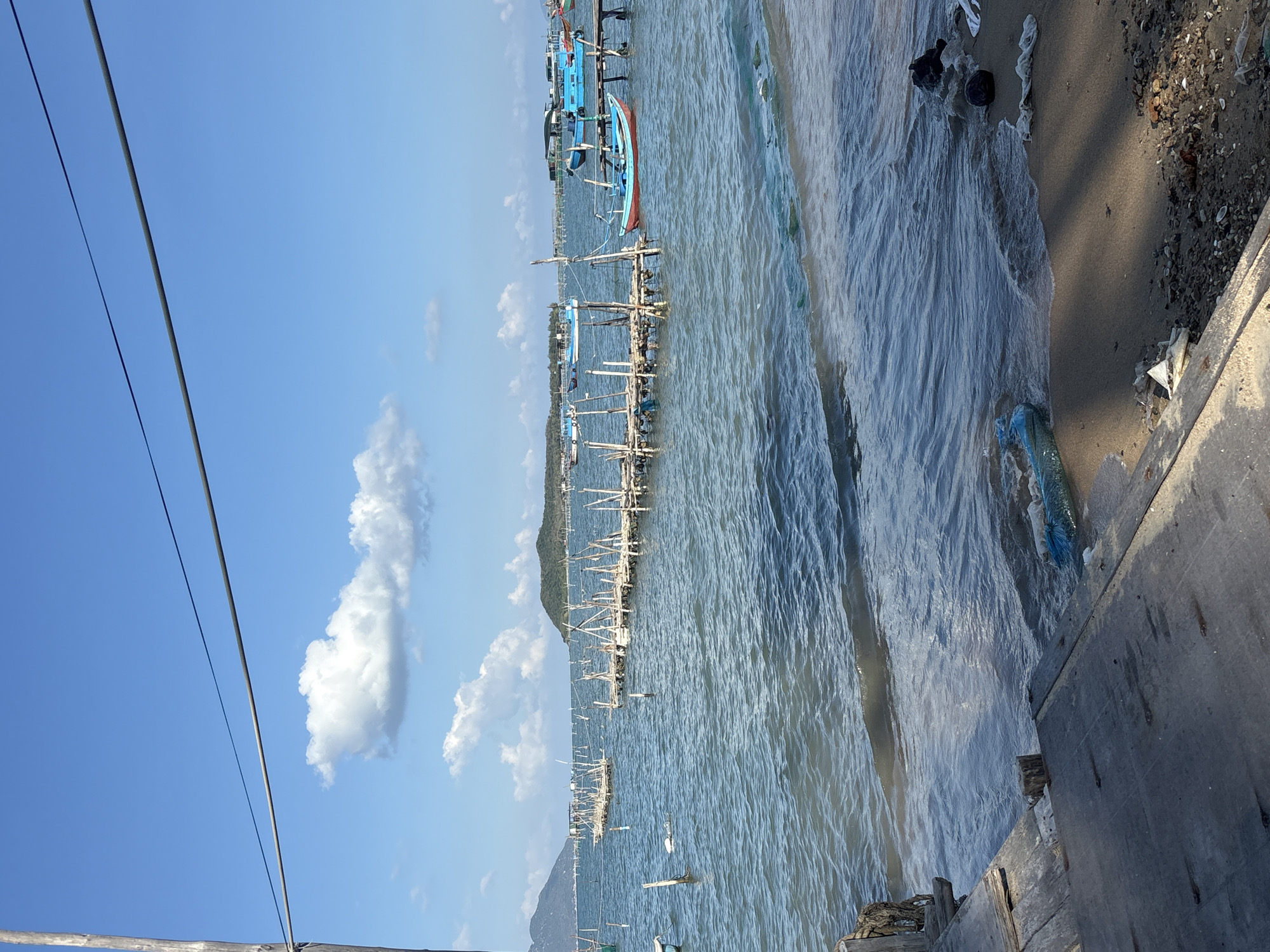 This image depicts a coastal scene in Bac Nha Trang Ward, Vietnam. The view is likely taken from an elevated position, possibly a pier or dock. The foreground shows weathered wooden planks. To the right, there's a sandy shoreline with some debris, including plastic waste. The main focus is the water, which has visible waves. Several fishing boats are docked in the water, primarily blue and red. A series of wooden structures, presumably for fishing, extend into the water. In the background, a hilly landscape is visible. The sky is blue with some scattered clouds. Overhead, there are electrical cables stretching across the sky.