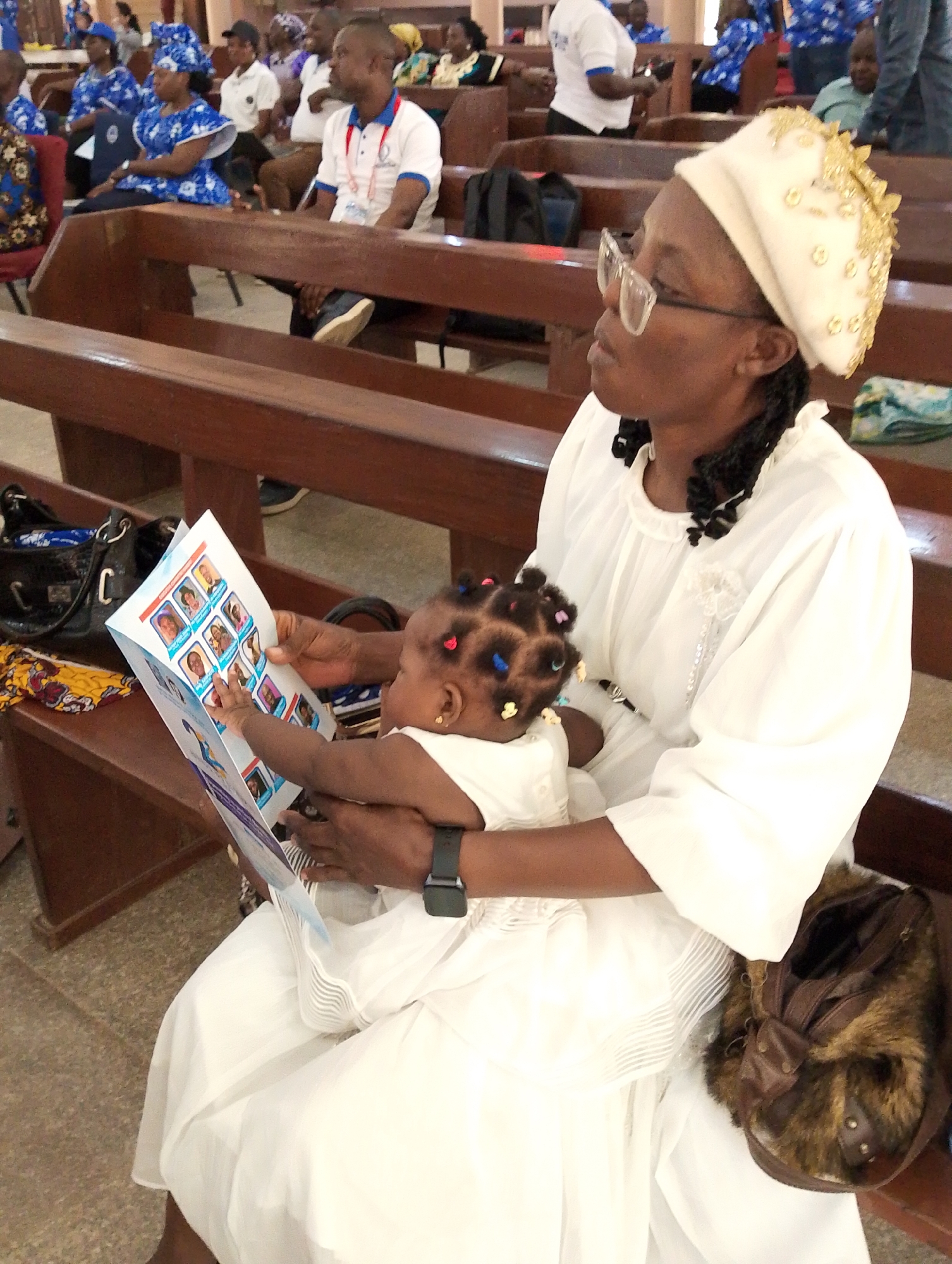 The image portrays an indoor scene within a hall or church in Kubwa, Nigeria, featuring a woman holding a baby on a wooden pew. The adult woman, dressed in a white, long-sleeved garment, wears a cream-colored head covering adorned with gold embellishments and clear-framed eyeglasses. Her hair is styled in dark braids.
She is attentively holding an open, folded pamphlet in front of the baby, who is seated on her lap. The baby, also in a white dress and sporting colorful hair ties, reaches out towards the pamphlet with a curious hand. The pamphlet displays a grid of multiple small portrait photographs on its pages. While the text beneath these images is too small to discern, it likely provides names or information related to the individuals pictured.
Beside the woman on the bench, a black handbag is visible, and a brown, fur-trimmed bag rests on her lap. In the background, numerous other individuals are seated on rows of similar wooden pews. Many attendees, including women in vibrant blue and white patterned traditional Nigerian attire, and men in white polo shirts with lanyards, are present. The lighting indicates it is daytime. The scene conveys a sense of calm and engagement, likely during a community gathering, church service, or a formal event.