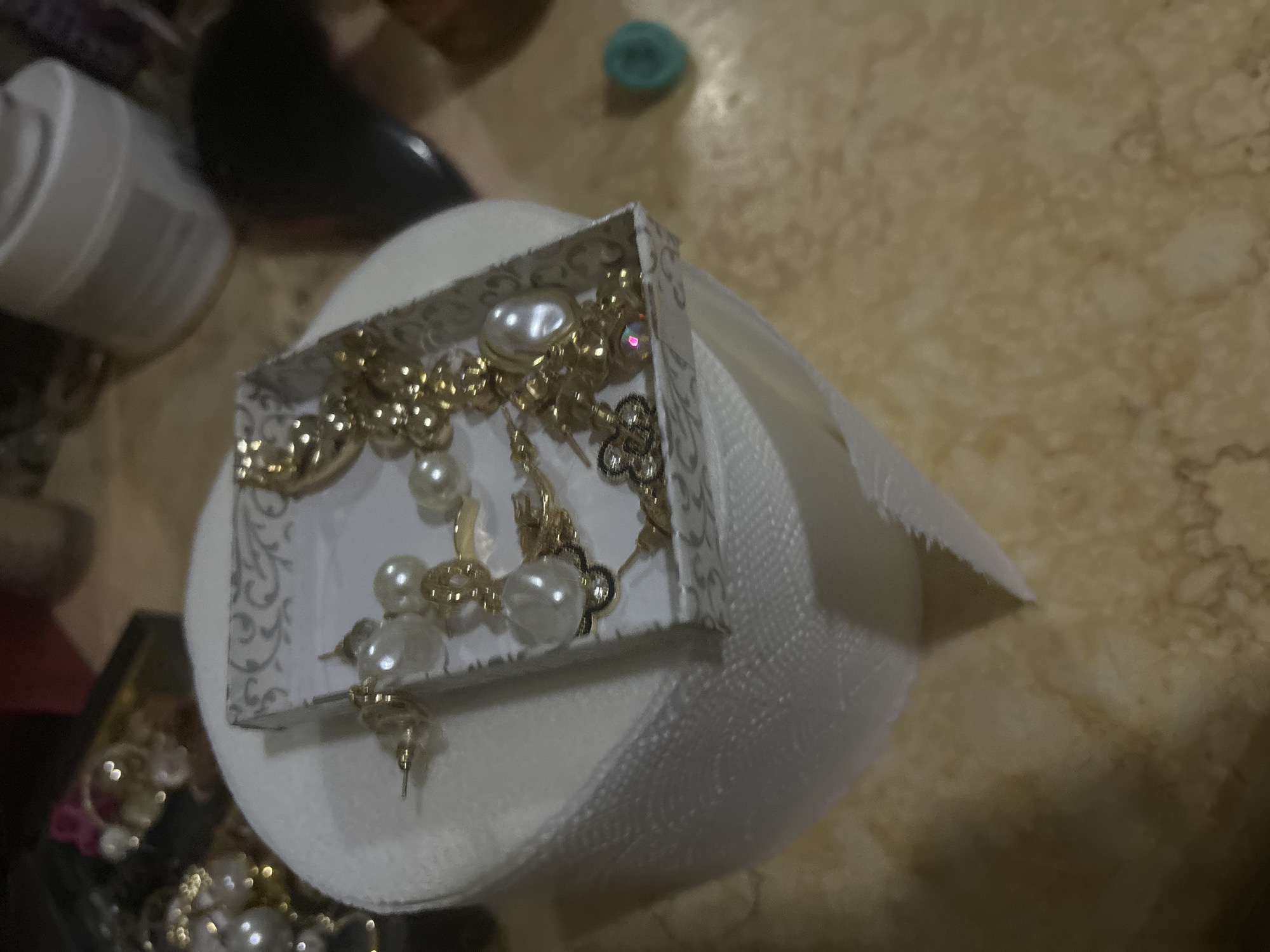 The image shows a small, white box filled with gold-colored jewelry, including pearl earrings and necklaces. The box is resting on a roll of toilet paper. The background includes a mottled beige surface, a white container, and some dark shapes. There is no information available that indicates the location of this image is in the city of Budva, Montenegro.