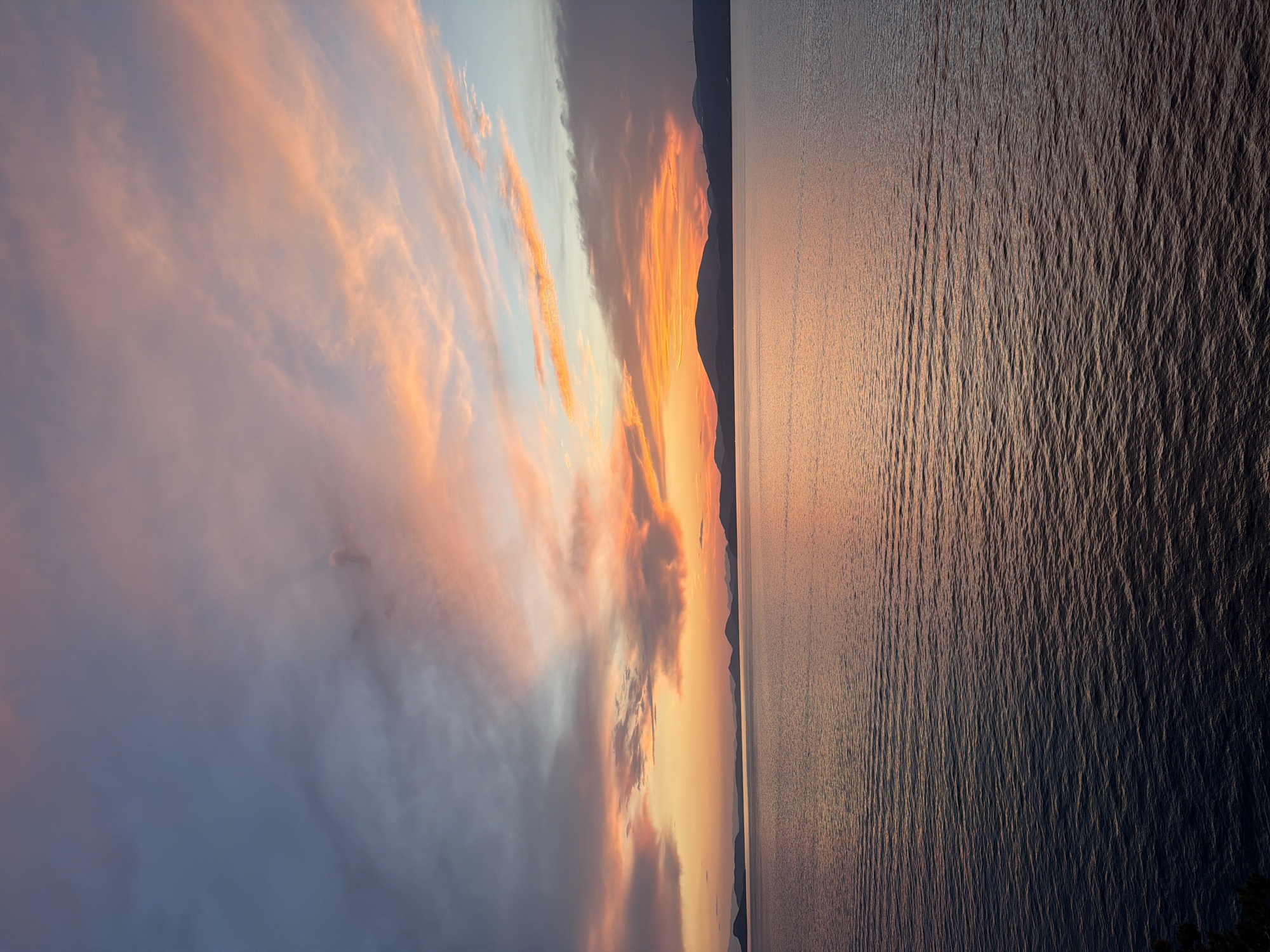 The image depicts a sunset view over the sea, with the silhouette of mountains or islands on the horizon. The sky is filled with clouds, painted with shades of orange, pink, and hints of blue. The sea reflects the colors of the sunset, displaying a dark, textured surface with visible ripples. The composition suggests the location might be near Hydra, Greece, given its mountainous landscape and island setting. There are no people or specific actions visible in the frame, focusing on the natural landscape and the colors of the sunset.