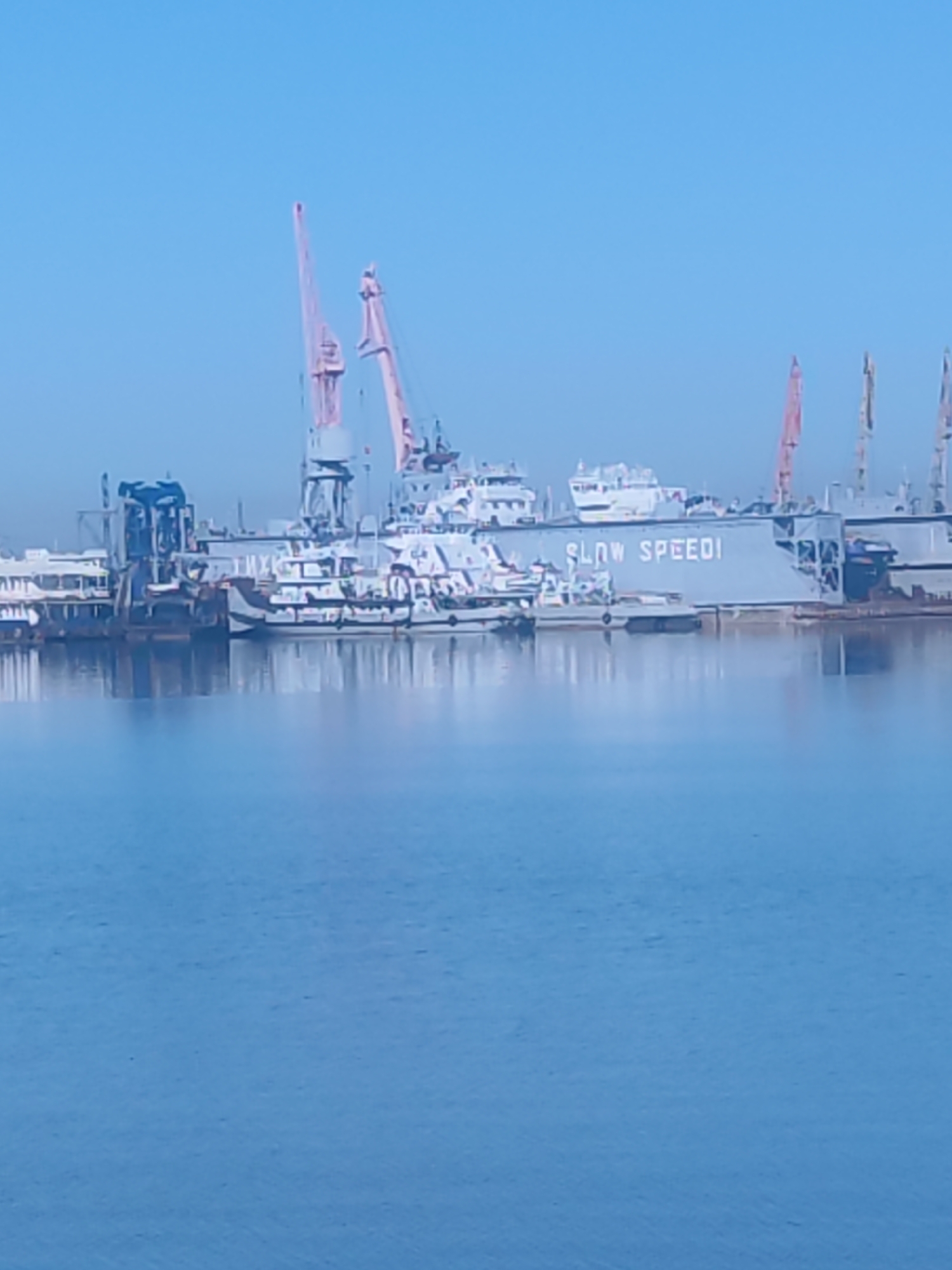 The image depicts a waterfront scene with various vessels and industrial structures against a clear blue sky. In the foreground, there is a large expanse of blue water. In the middle ground, there's a dock area with multiple ships of different sizes. Cranes with pink booms are visible, suggesting a shipyard or port facility. One of the larger gray-colored vessels has the words "SLOW SPEED!" painted on its side in white lettering. There are also smaller boats docked along the pier. The location is likely a port city in the city of Unknown, Unknown.