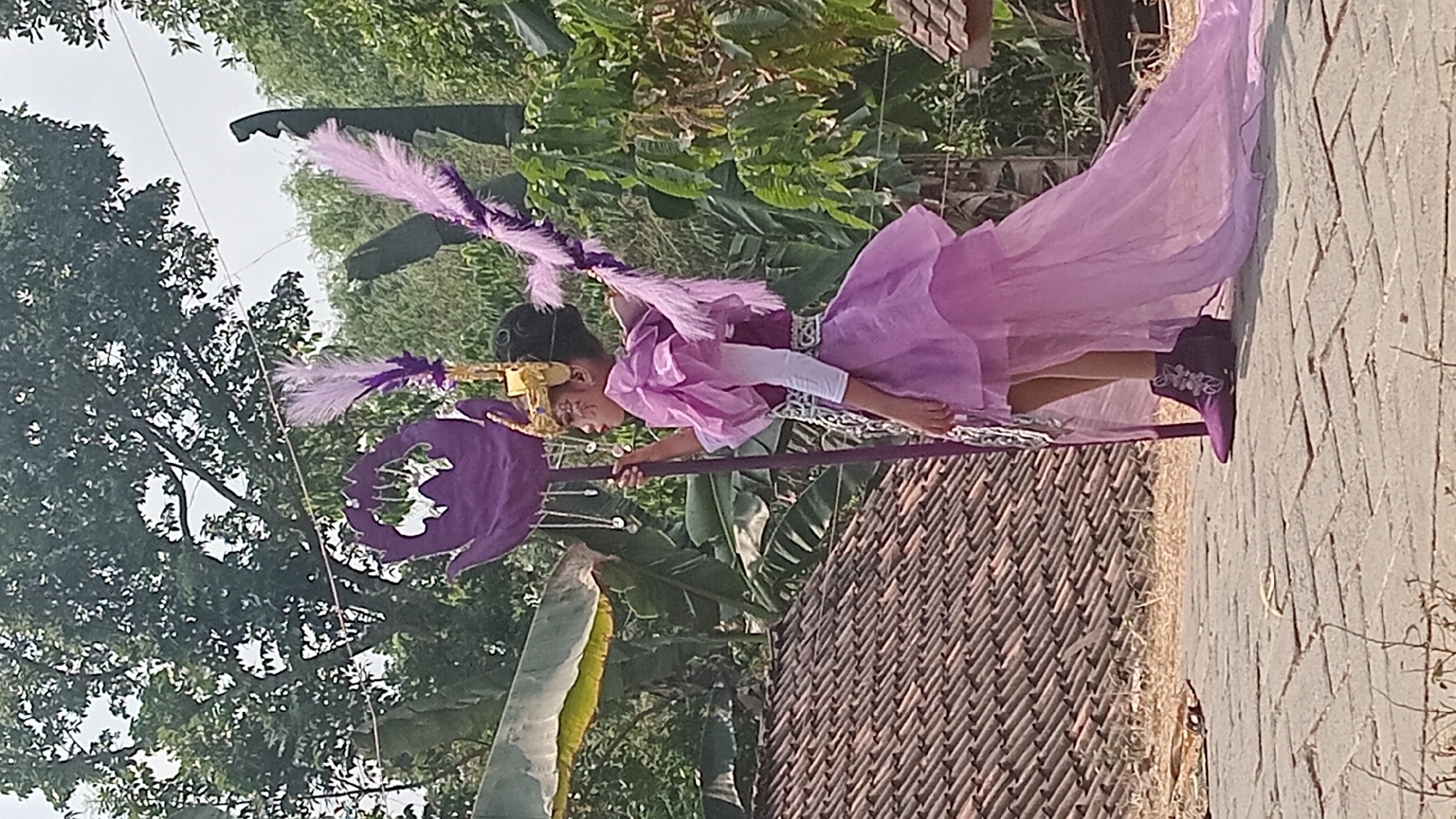 An Indonesian child, likely female given the attire, stands on a paved surface in Kedungwaru, Indonesia. The child is dressed in a theatrical, light purple costume with puffed sleeves, a layered skirt, and a long train. A gold headdress with feather accents adorns the child's head, and ornate makeup is visible. The child holds a tall, purple staff topped with a lotus-like design and adorned with hanging crystals. Large green leaves and trees form the background, with a tiled roof of a building visible to the left and a wooden structure to the right. The lighting suggests it is daytime, with clear skies.