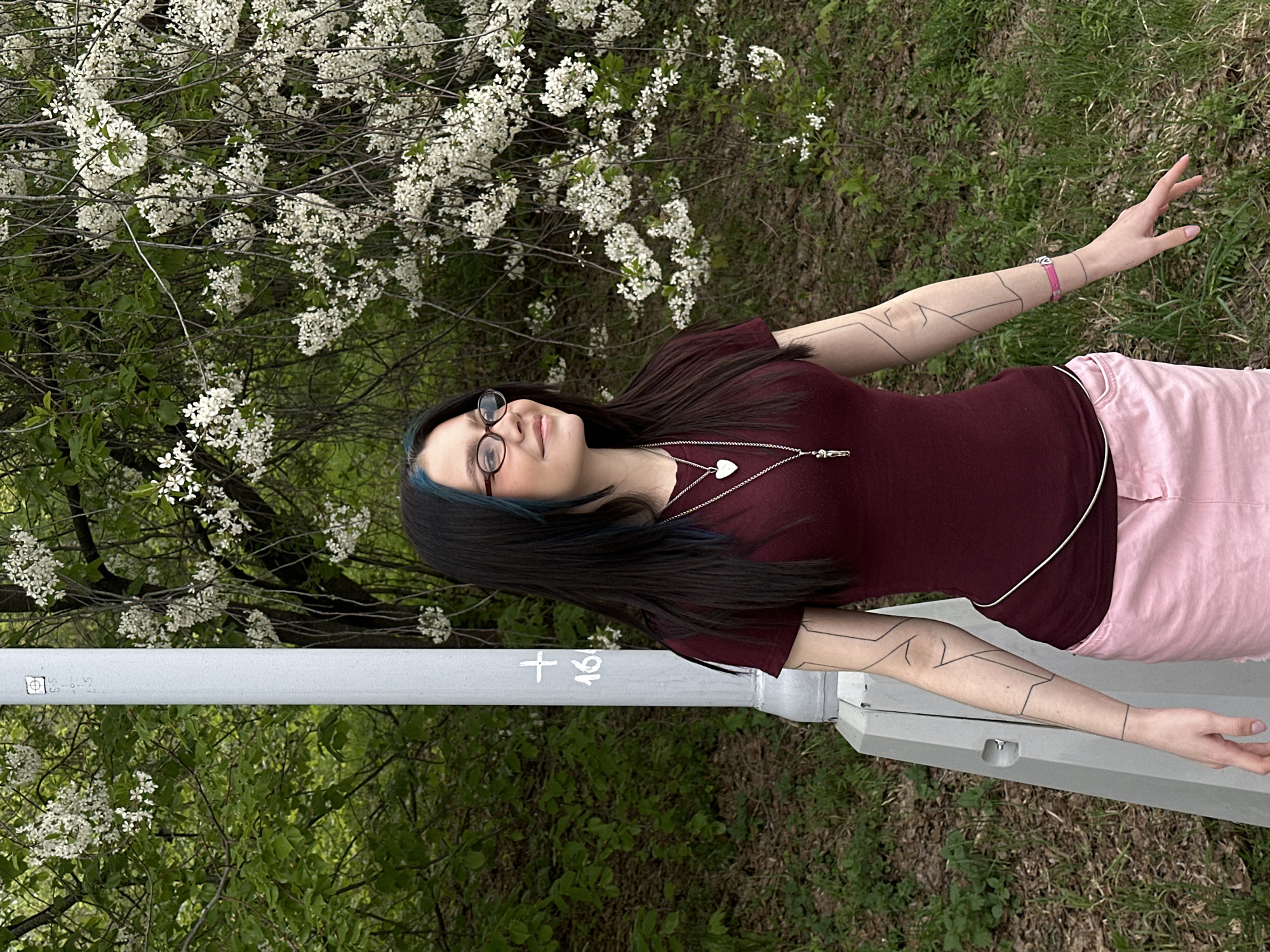 A young woman stands outdoors next to a light pole in a grassy area with trees and blooming white flowers. She is wearing glasses, a maroon t-shirt, and pink pants. She has black hair with a streak of blue and a silver necklace with a heart pendant. Her arms are outstretched, and she has geometric designs drawn on her forearms. The light pole has numbers and symbols on it. Given the flora and architectural style, the location might be in Moscow, Russia, but this is only a guess based on the visual context.