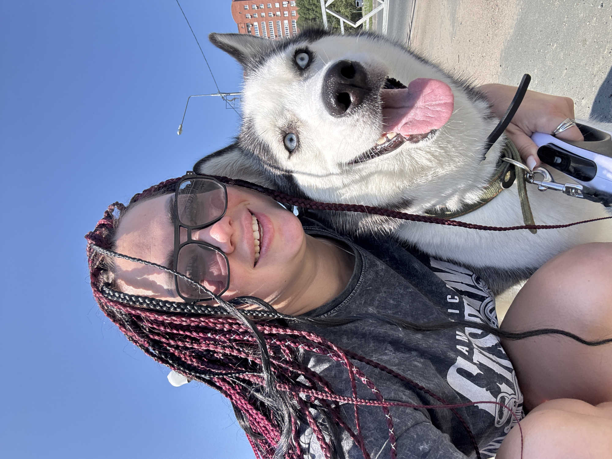 In Tolyatti, Russia, a young woman with dark braided hair, accented with burgundy strands and secured with white string, takes a selfie with a Siberian husky. She wears large, dark-framed sunglasses and a grey, acid-wash t-shirt that displays the text "AMERICAN CLASSIC." The husky, positioned to her right, has striking blue eyes, a pink tongue hanging out, and a black and white coat. The woman's left hand is visible, holding a white retractable dog leash, and she wears multiple silver rings. They are outdoors, with a bright blue sky overhead. In the background, a street lamp and a multi-story brick building are visible, along with trees and a bridge-like structure, suggesting an urban setting. The ground appears to be paved.