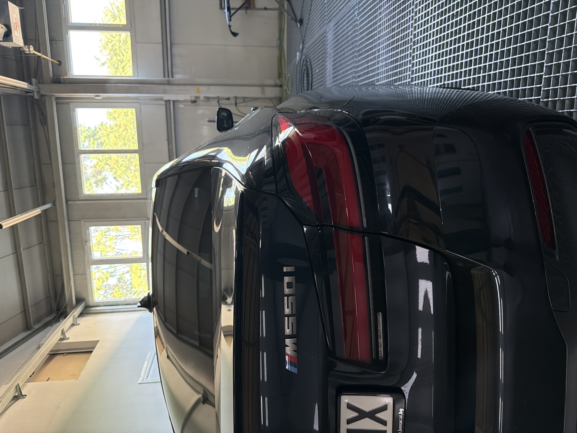 The image shows the rear passenger side of a black BMW M550i parked inside a garage or workshop. The car's taillight and trunk lid are visible, with the "M550i" badge prominently displayed. The interior of the garage features white walls, a metal grating floor, and several windows with sunlight streaming through, illuminating trees outside. A license plate with the letters "IX" is affixed to the rear of the car, and part of a website address "www.bovacar.bg" is visible below it, indicating the location is likely in Burgas, Bulgaria, as Bovacar is a car dealership located in Burgas. There are no people visible in the image.