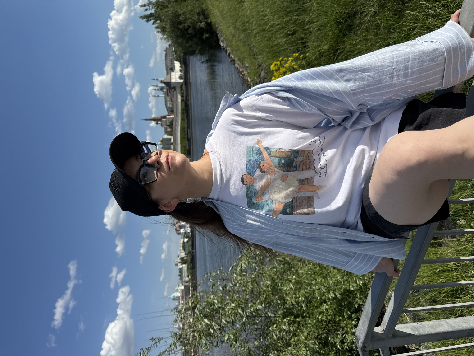 A person wearing sunglasses, a baseball cap, a t-shirt with an image of people dancing, and a light blue button-down shirt is sitting on a metal railing. The scene appears to be outdoors on a sunny day. The background includes a body of water, greenery, and a cityscape featuring recognizable buildings and structures of Kazan, Russia.