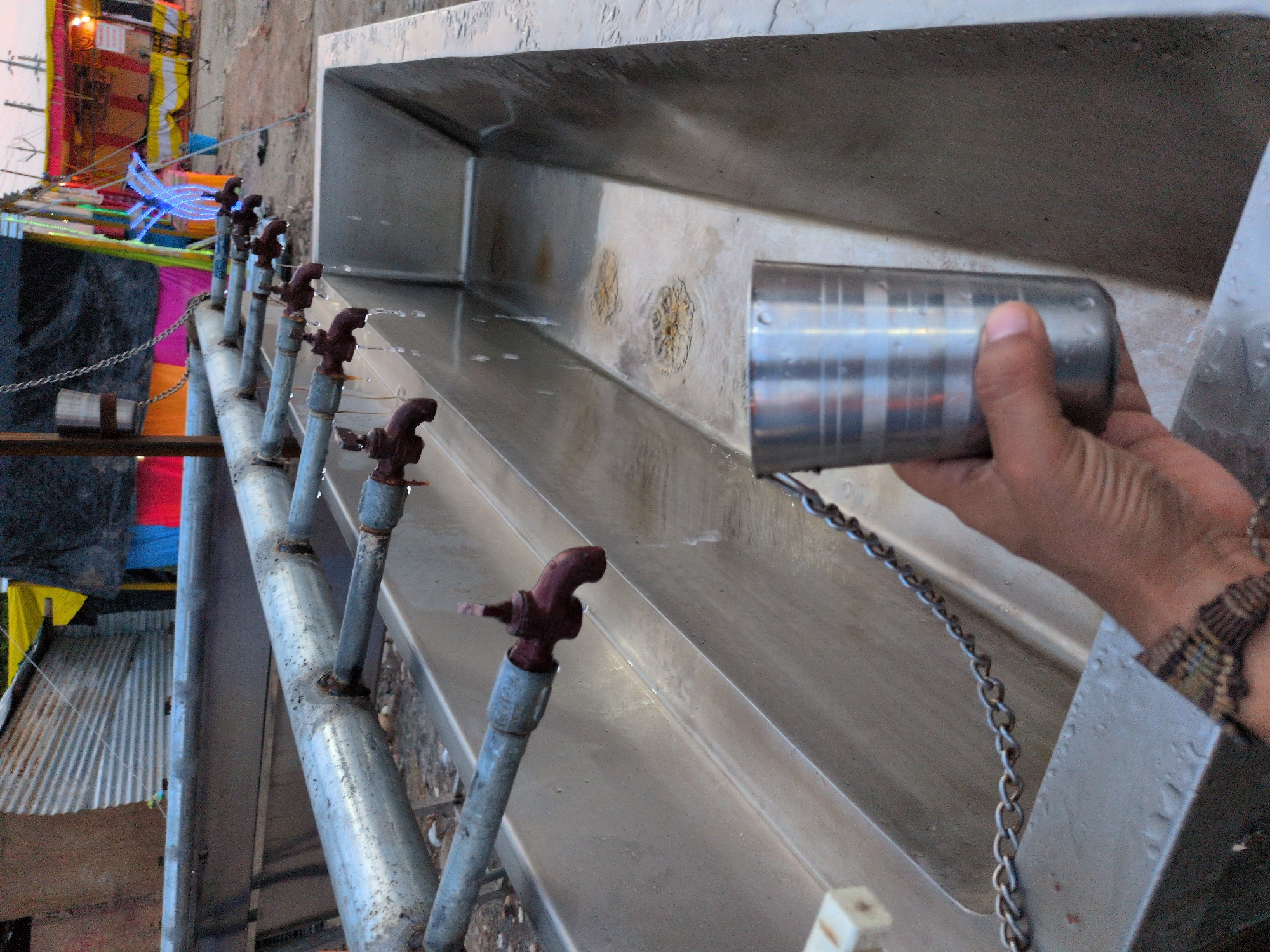 A man's hand holds a metallic cup under a row of outdoor water taps. The taps are attached to a silver pipe running parallel to a large, shallow metallic trough. Water is dripping from several of the taps into the trough. The trough is partially filled with water, and there are circular, stained patterns on the bottom. A silver chain is visible attached to the trough and extends towards the left of the frame. In the background, there are temporary structures with colorful fabric awnings and signage, typical of a festival or event setting. The ground appears to be unpaved. The overall impression is a public water dispensing area, possibly at a gathering or market. The scene is likely in Jammu, India, given the context.