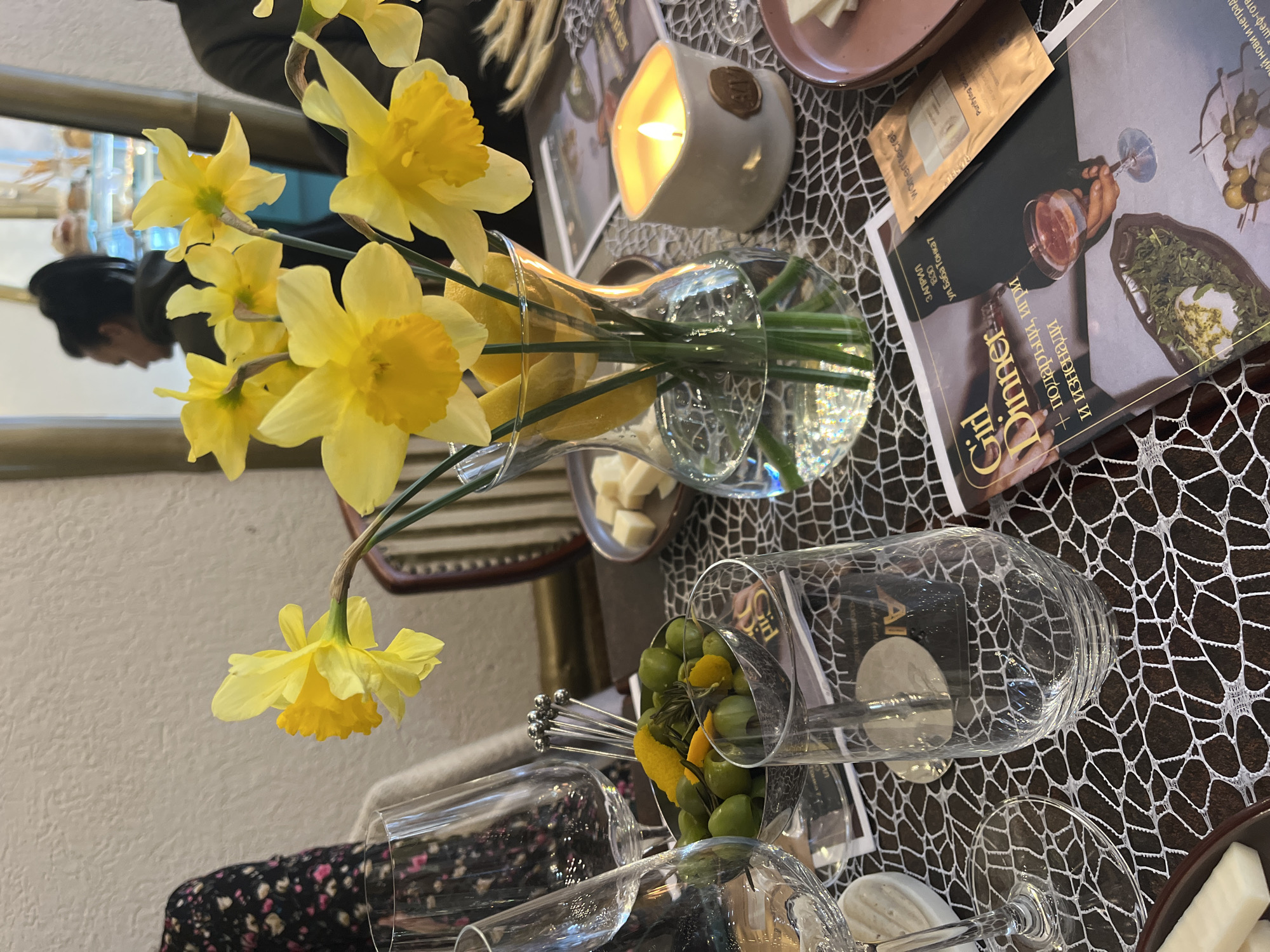 The image shows a table setting for a "Girl Dinner" event in Varna, Bulgaria. The table has a black and white patterned tablecloth. A glass vase holds yellow daffodils with lemons. There is a lit candle, a plate of cheese, a small packet of cosmetics, and a book titled "Girl Dinner + подаръци, игри и изненади" (translated as "Girl Dinner + gifts, games and surprises"). Several wine glasses are lying on their sides, with one containing green olives and lemon slices. A small set of cocktail forks are also present. A reflection of a person can be seen in a mirror in the background. The location is given as ул. Баба Тонка 7 (7 Baba Tonka Street).