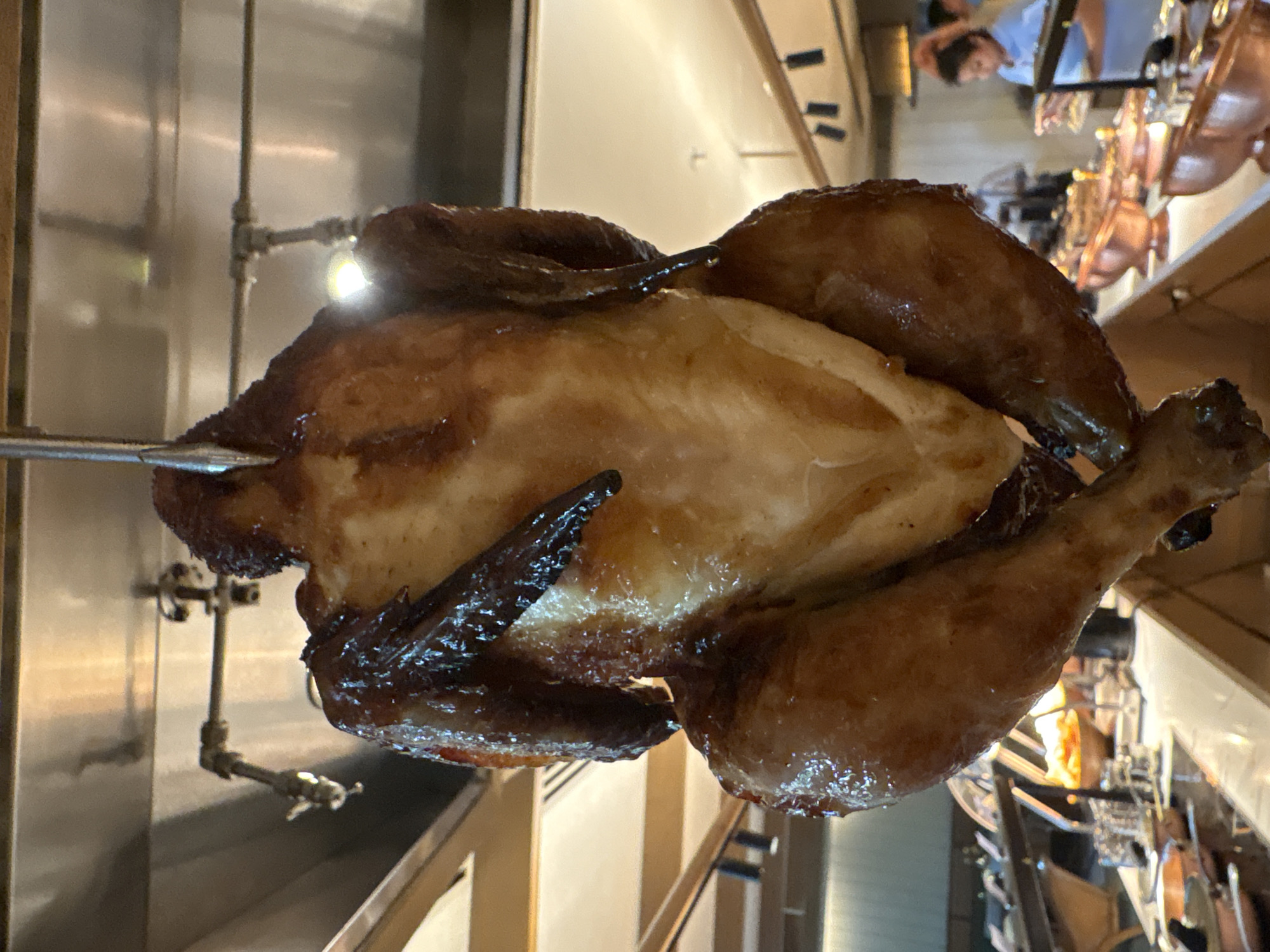 A whole roasted chicken hangs from a metal skewer, its skin a glistening golden brown. The chicken is suspended in front of a blurred background that appears to be a buffet or food service area. Visible elements in the background include buffet chafing dishes, some containing food, and a person in a blue shirt standing behind a counter, possibly a food service worker. The lighting is warm and ambient, suggesting an indoor setting, possibly a restaurant or hotel. The composition focuses on the chicken, which is well-lit and appears appetizing. The image does not provide specific details to locate it in Vagator, India.