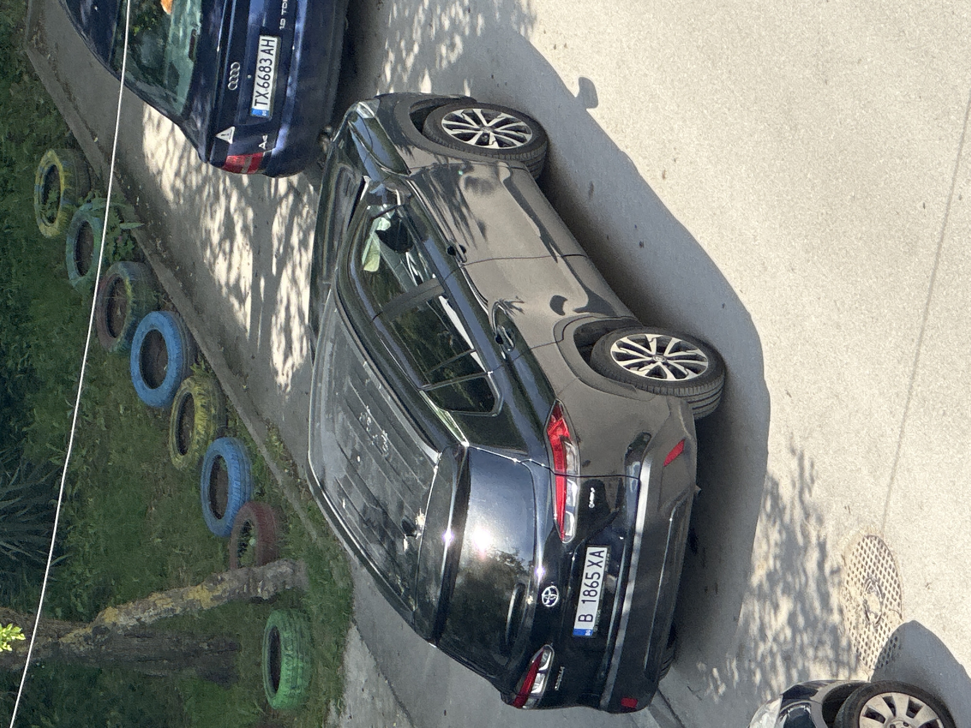 The image depicts a street scene in Varna, Bulgaria. A dark gray Toyota RAV4 with license plate B 1865 XA is parked parallel to the curb. To the left of the vehicle is a line of painted tires decorating the edge of a grassy area. A blue Audi A4 with license plate TX 6683 AH is parked in front of the Toyota. The street is paved and shows signs of wear. A manhole cover is visible on the right edge of the frame.