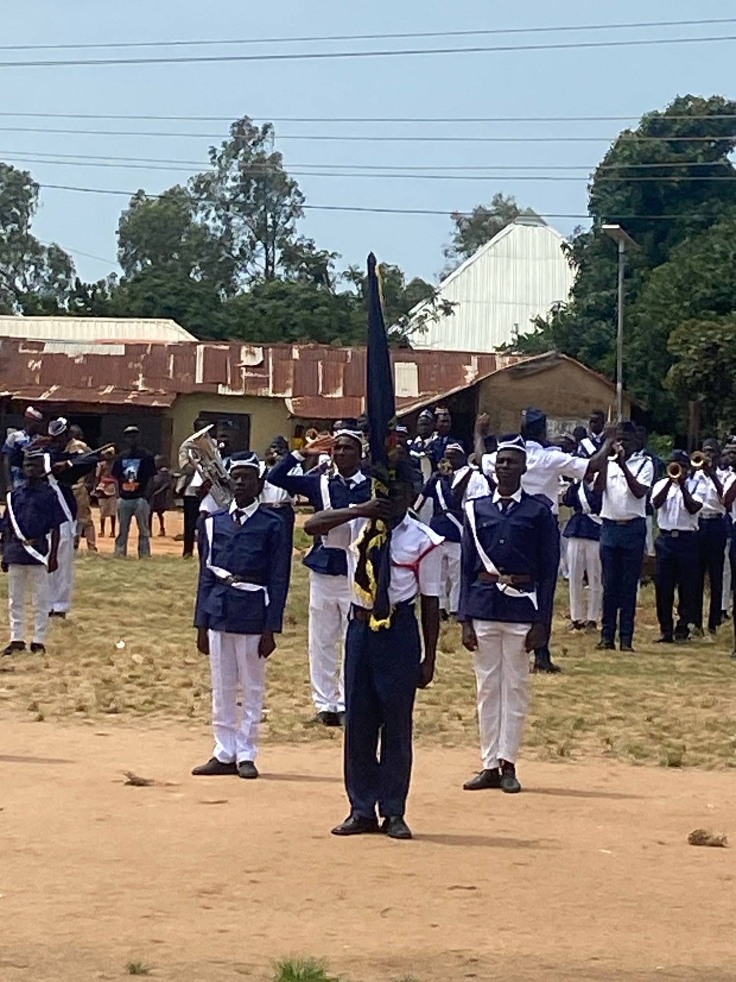 In Binchi, Nigeria, a group of uniformed individuals stand in formation on a dirt field. The individuals are wearing dark blue jackets with white trim and white pants, and some have white sashes. Many are wearing white caps with dark bands. In the foreground, one individual holds a large dark flag with yellow trim. Several individuals in the background are holding musical instruments, including trumpets and a tuba. Behind the formation, there are rustic buildings with corrugated metal roofs, and some trees and power lines are visible against a pale sky. The ground is dry and dusty, with sparse patches of dry grass.