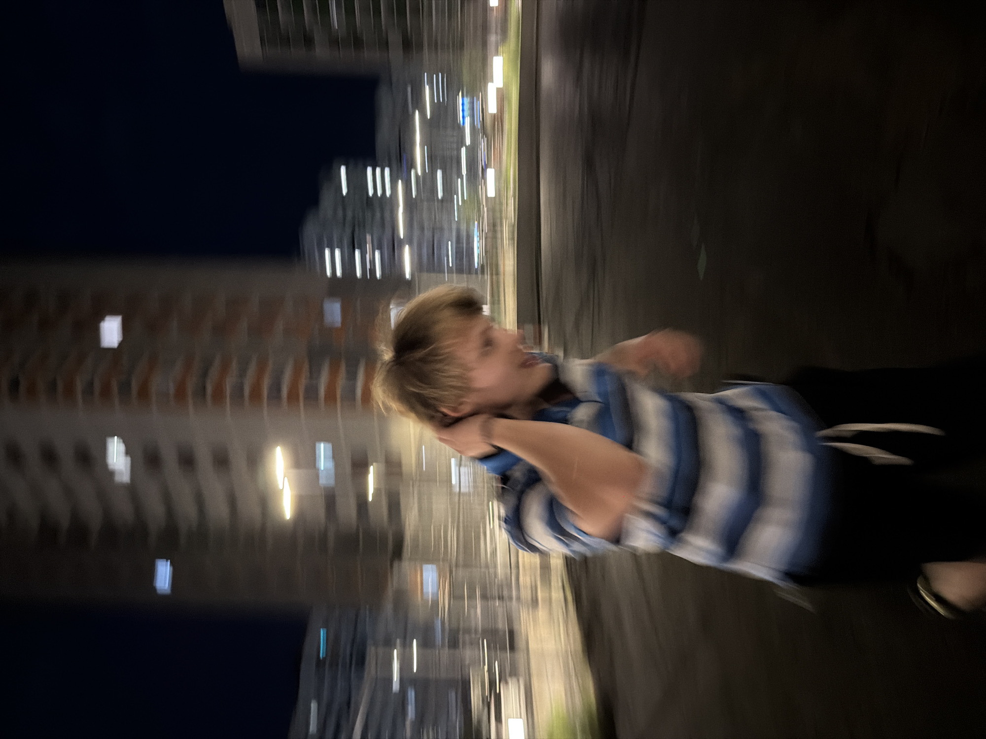 The image depicts a young man with blonde hair, wearing a blue and white striped polo shirt and black shorts, in motion at night. He is running or moving rapidly, with his right arm raised and touching the back of his neck. The background shows blurred elements of an urban environment at night, including tall buildings with illuminated windows and streetlights, suggesting he is in a city. The lighting is low, and the motion blur indicates movement. The scene is consistent with a residential or commercial area in Izhevsk, Russia, during nighttime.