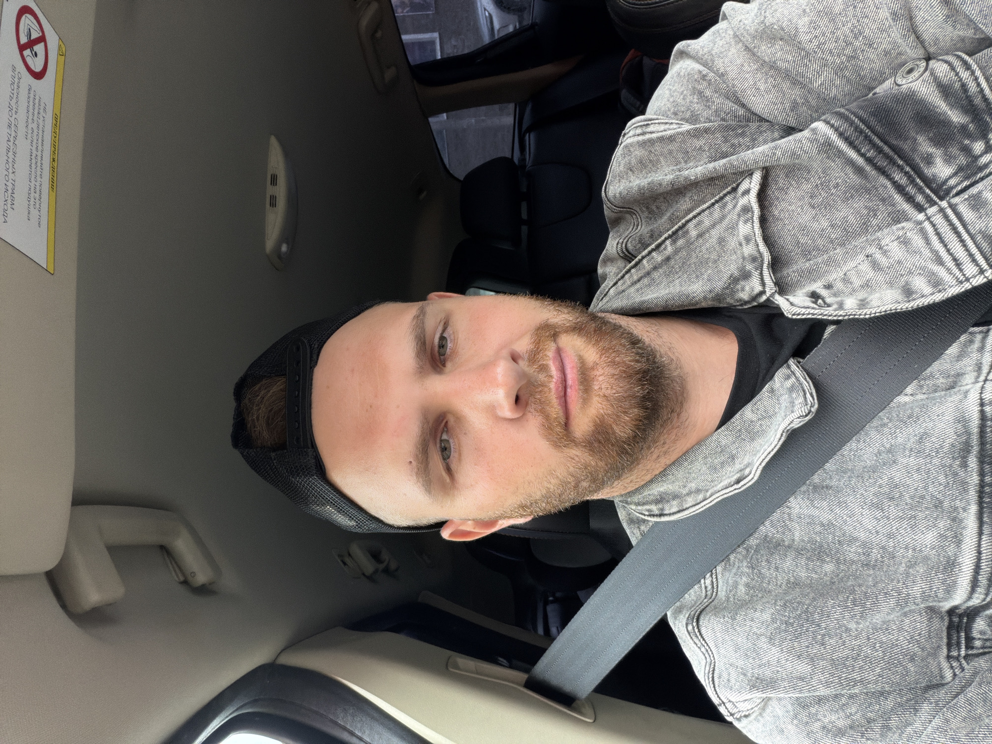 An adult male with a reddish-brown beard and short brown hair, wearing a black mesh cap backward, is seated inside a vehicle. He is wearing a washed grey denim jacket over a black garment, with a dark grey seatbelt secured across his chest and shoulder. He looks directly at the camera.
The car's interior features a light-colored headliner, an overhead grab handle, and an overhead light/microphone unit. A yellow warning label with Cyrillic text is visible on the passenger-side sun visor. The rear seats are upholstered in dark material. Through the tinted rear windows, out-of-focus brick or concrete buildings consistent with an urban environment, and part of another light-colored vehicle, are discernible. The scene is illuminated by daylight, suggesting an overcast sky or shaded conditions.