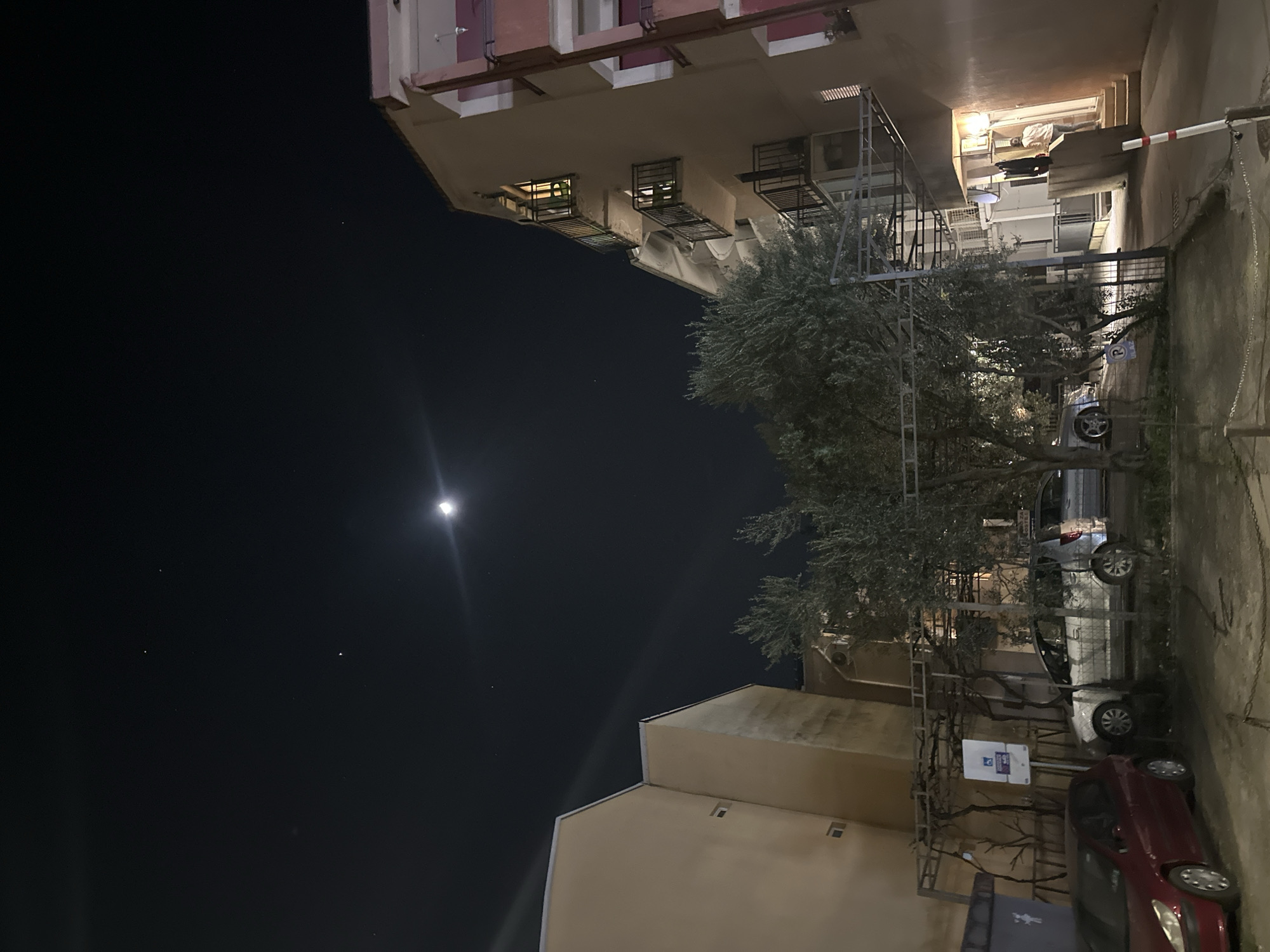The image depicts a nighttime scene in a residential area of Budva, Montenegro. A crescent moon is visible in the upper right quadrant of the dark sky, accompanied by faint stars and a lens flare emanating from the moon.
The scene is dominated by multi-story residential buildings. On the left, a light-beige building with a plain facade and small rectangular windows is visible. On the right, a multi-story building with a light-beige and pink/reddish-brown facade features multiple balconies with black metal railings; some interior lights are illuminated. An air conditioning unit is mounted on the exterior wall of this building.
In the midground, a mature olive tree with dense foliage is prominent, partially obscuring a silver hatchback and another vehicle behind it. A metal trellis structure stands near the tree, stretching towards the left.
The foreground consists of a loosely paved or unpaved ground surface. Parked vehicles include a red hatchback on the far left, adjacent to a dark grey dumpster. A blue and white sign with the international handicapped symbol is mounted near the center-left. A chain barrier is visible across the lower foreground.
On the far right, near an illuminated doorway, a single individual is observed from behind, wearing a light-colored top and dark trousers. They appear to be near the entrance or exit of the building. A white and red striped bollard or marker is positioned in the lower right foreground.