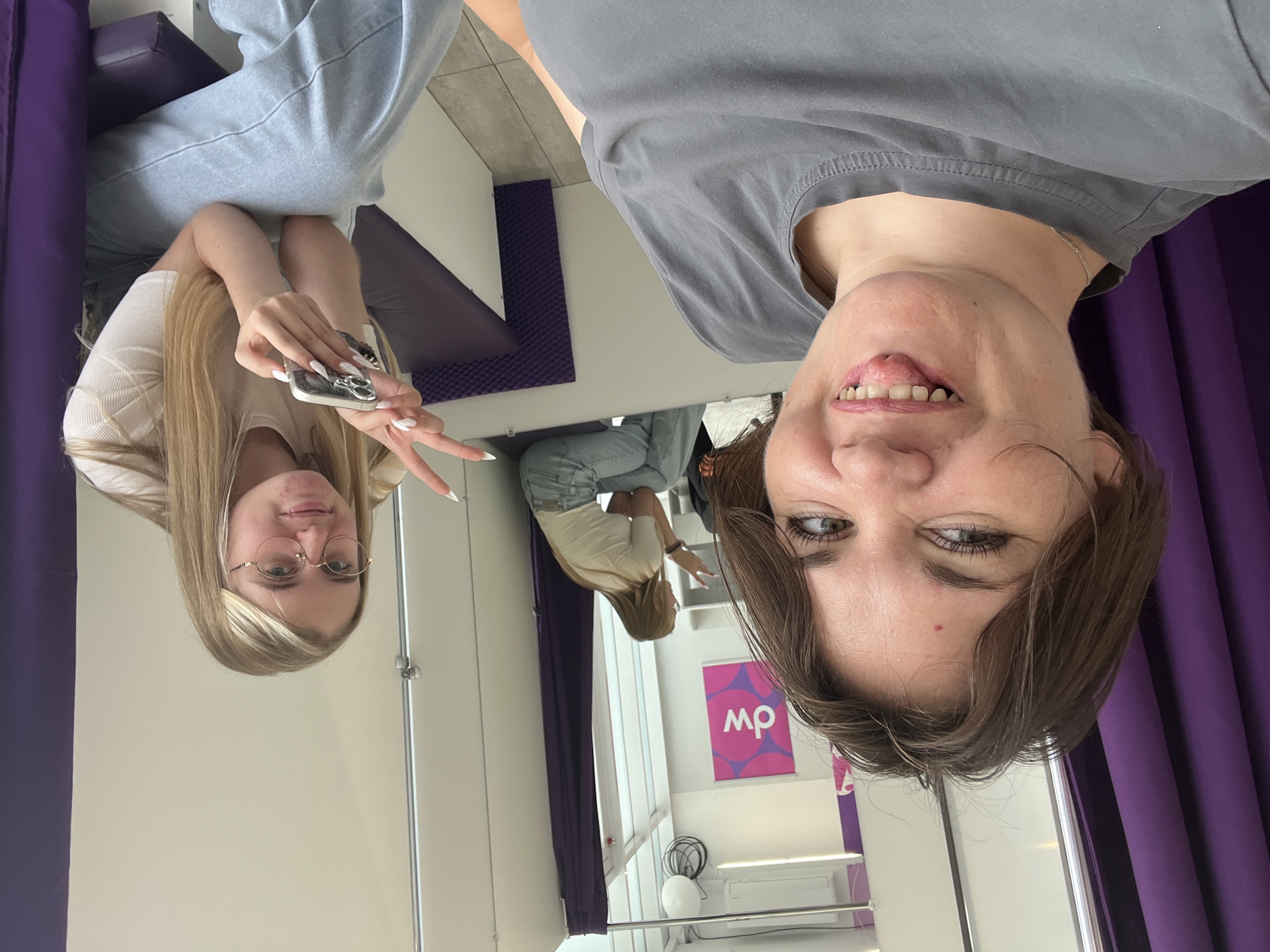 Two young women are inside a changing room, likely at a retail store. One woman, in the foreground and to the left, is taking a selfie. She has short brown hair, is wearing a grey t-shirt, and is sticking her tongue out at the camera. The second woman is seated to the right, wearing a white t-shirt and light blue jeans, and is holding a smartphone and making a peace sign with her fingers. A mirror behind them reflects the interior of the changing room, including a pink and purple "dw" logo on the wall. The changing room has purple curtains, white walls, and a grey floor. The location is identified as Afipskiy, Russia.