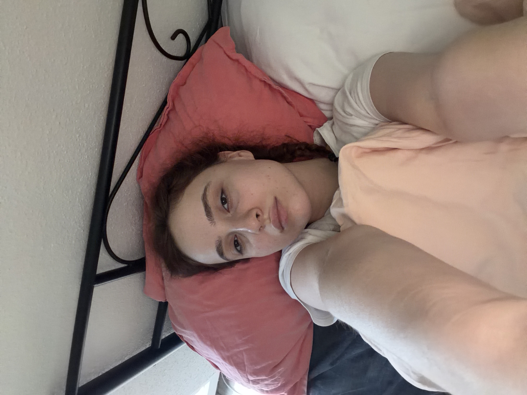 An individual, a young female, is depicted lying in a bed in an indoor setting. Her head rests on a large, rumpled coral-pink pillow positioned against a dark gray sheet. A portion of a white pillow is visible to her right. The individual has light skin, dark brown eyes, and dark reddish-brown hair, with a visible braid along her left side. She wears a white short-sleeved top and is partially covered by a light pink blanket or duvet that extends over her torso. Her right arm, bent at the elbow, is partially visible. Her left arm is mostly obscured. She maintains a neutral facial expression, looking directly towards the camera. Above her head, a black metal headboard with a decorative scroll is present. The background reveals a textured, off-white ceiling or wall. This scene is located in Strasbourg, France.