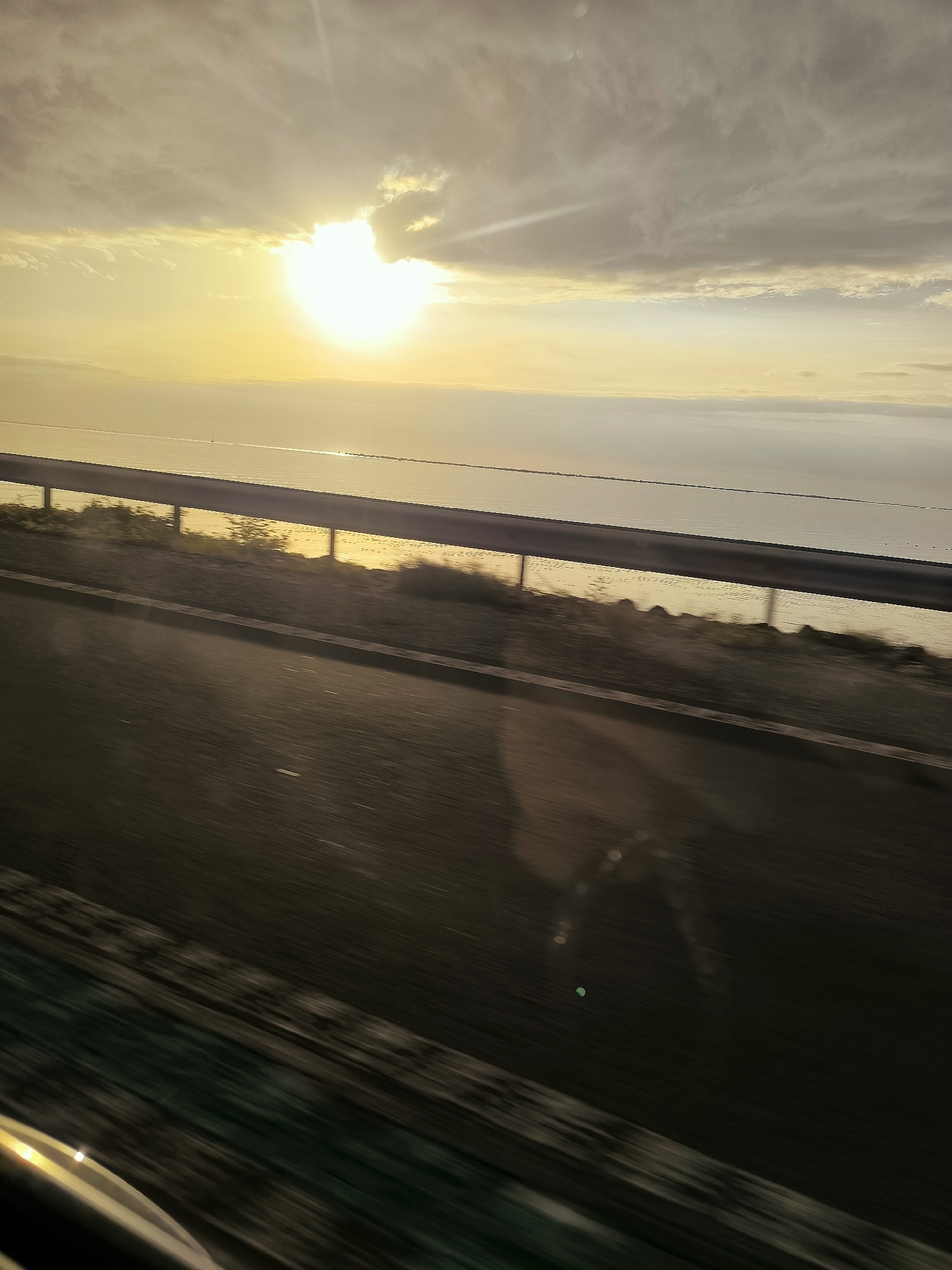 The image depicts a sunset over a body of water, likely the IJsselmeer, viewed from a road with a guardrail. The sky is filled with clouds, and the sun is partially obscured by one. The water reflects the light of the setting sun. A road is visible in the foreground, with a guardrail separating it from the water. The environment appears to be flat and open, typical of the Flevopolder region. The image seems to have been taken while in motion, possibly from a vehicle, as indicated by the motion blur on the road and the surrounding vegetation. There are no visible people or specific buildings that would pinpoint the exact location within Lelystad.