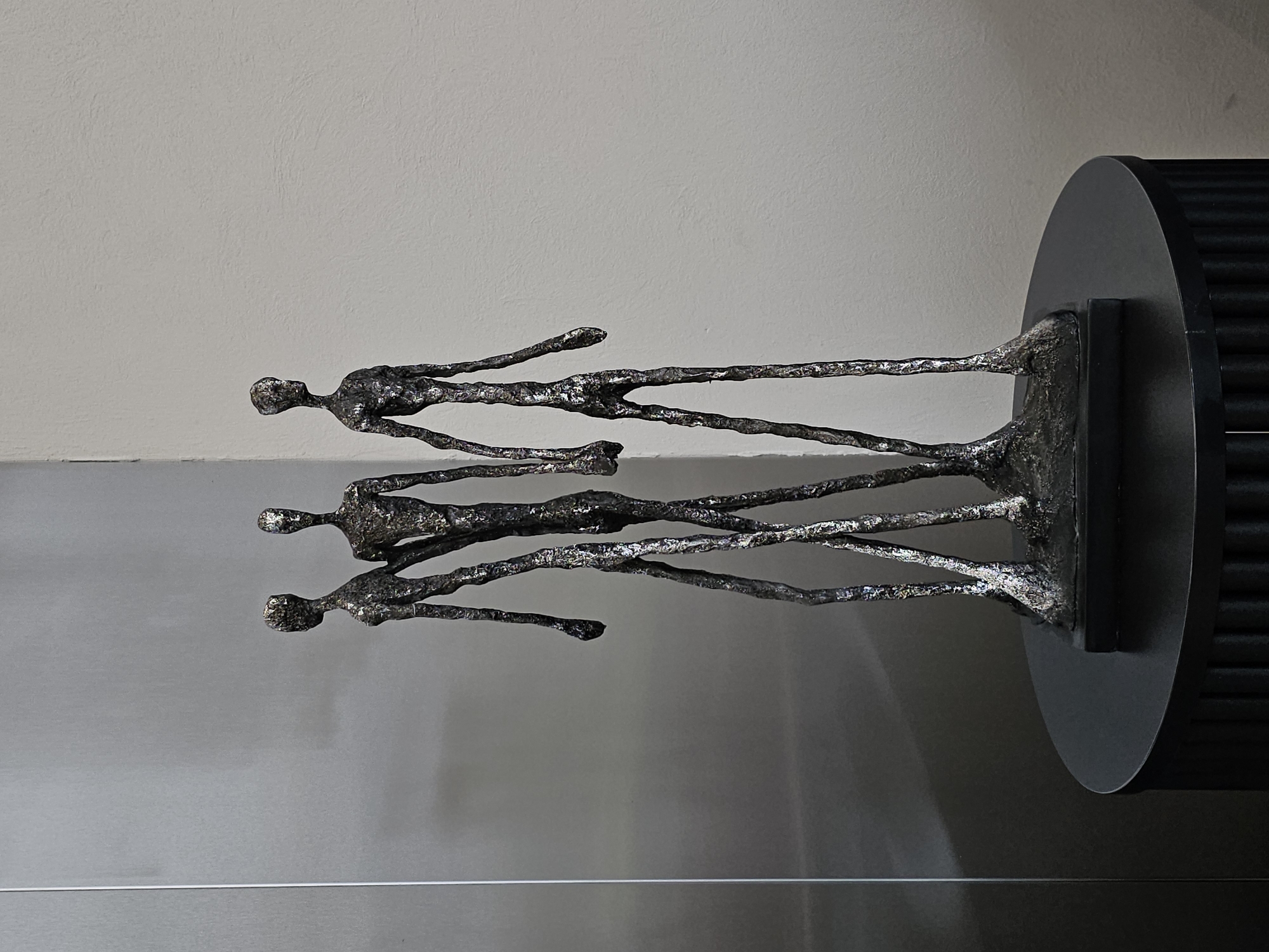 A metal sculpture of three elongated, stick-like figures stands on a black pedestal. The figures are dark and textured, appearing to be made of bronze or a similar metal with a patina. They are arranged close together, with one figure slightly behind and between the other two. The sculpture is placed on a circular black table with a fluted base. The background consists of a smooth, light gray wall on the right and a reflective, metallic surface on the left. The lighting is soft, casting subtle shadows. The scene appears to be indoors, possibly in a gallery or a modern interior space. There are no people visible in the image, and no specific location in Moscow, Russia, can be determined from the visual information provided.