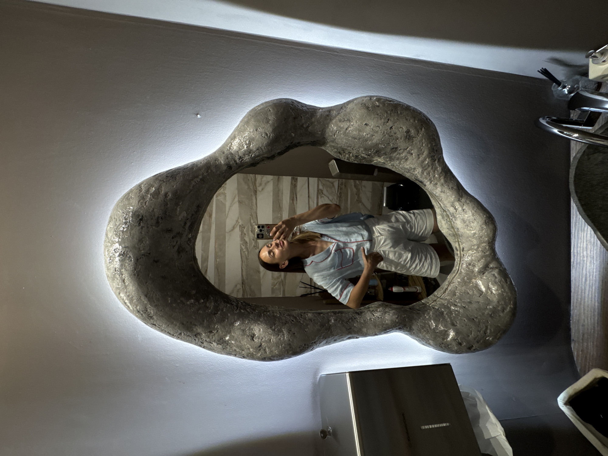 A woman in pajamas takes a mirror selfie in a bathroom. She is wearing a light blue button-up shirt with red piping and white shorts. She is standing in front of a large, irregularly shaped mirror with a textured, gray frame that is illuminated from behind by LED lights. In the reflection, the bathroom features marble-patterned walls and various toiletries on shelves. To the left of the mirror, a stainless steel dispenser is mounted on the wall. The environment appears to be a modern bathroom, possibly in a hotel or residential setting. The image does not provide specific details about the city of Sochi, Russia.