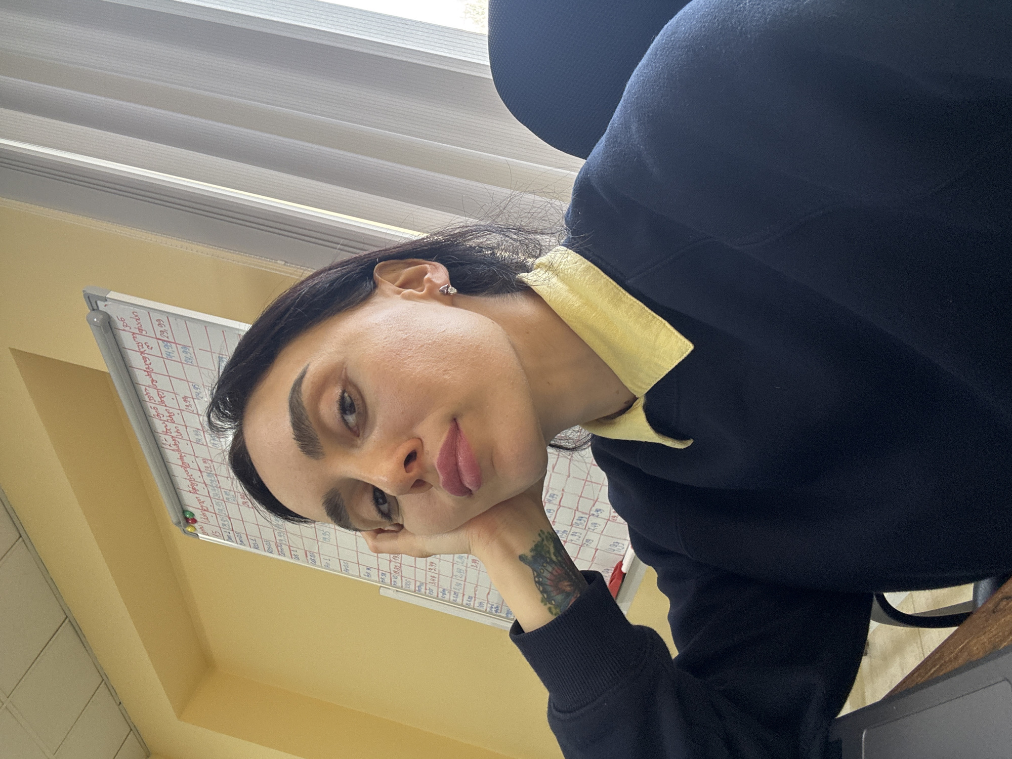 A medium shot shows a person with dark hair and eyebrows, leaning their head on their left hand, which has a visible butterfly tattoo on the wrist. The person is wearing a dark blue or black sweatshirt with a yellow collar. The background features a whiteboard with handwritten text and numbers in red and blue, along with a window with horizontal blinds. The room has a yellow wall. Although it is impossible to pinpoint the exact location in Tbilisi, Georgia, based on the image alone, the indoor setting suggests an office or a study room.