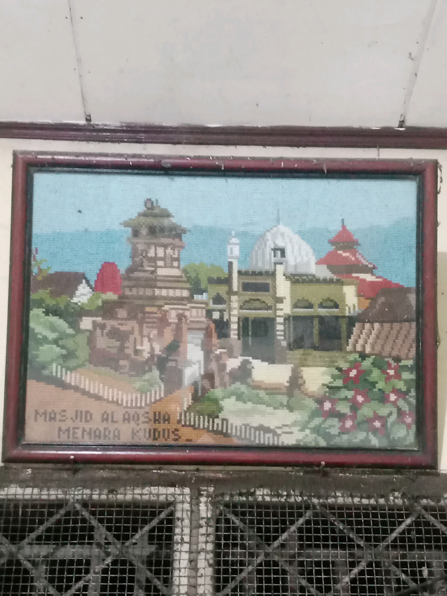 The image shows a framed tapestry depicting the Masjid Al-Aqsha Menara Kudus, a mosque in Kudus, Indonesia, with its distinctive tower. The tapestry is in a pixelated, cross-stitch style and features a light blue sky and a landscape with buildings, trees, and red flowers. The text "MASJID AL-AQSHA MENARA KUDUS" is stitched at the bottom of the tapestry. The framed tapestry is hanging on a light-colored wall above a metal grate. The wall appears to have some dust or debris on it. The overall scene suggests an indoor setting, possibly a home or a place of worship, with the tapestry serving as a decorative element. The provided context mentioning Balaraja, Indonesia, is not visually supported by the image itself.