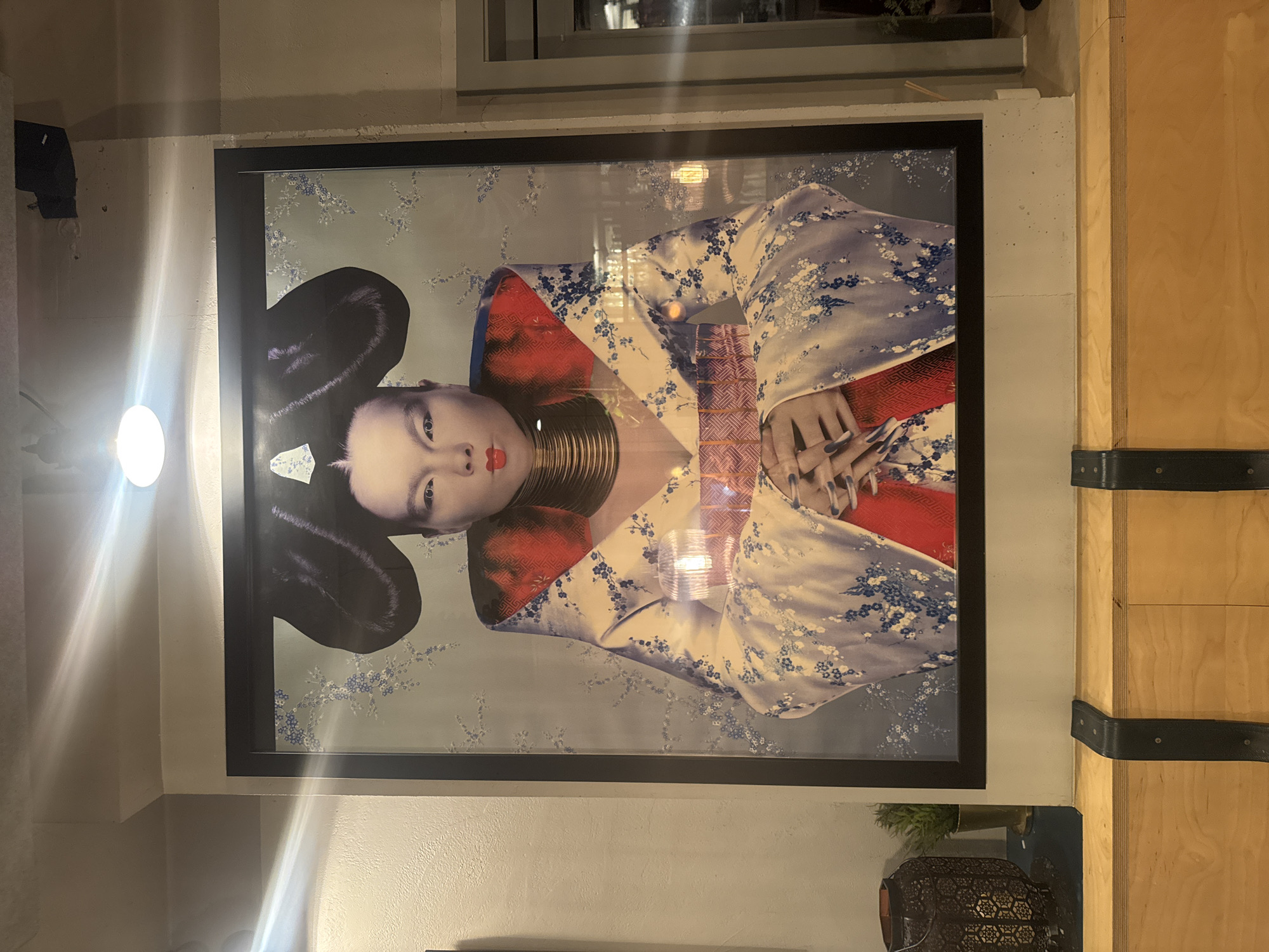 The image shows a framed portrait of a woman in a traditional Japanese-style outfit, with elaborate hair, neck rings, and a patterned kimono, hanging on a wall next to a wooden structure secured with black straps. The photograph is well-lit, but there is a glare that interferes with the picture. The photograph is located in a building in the city of London, United Kingdom.