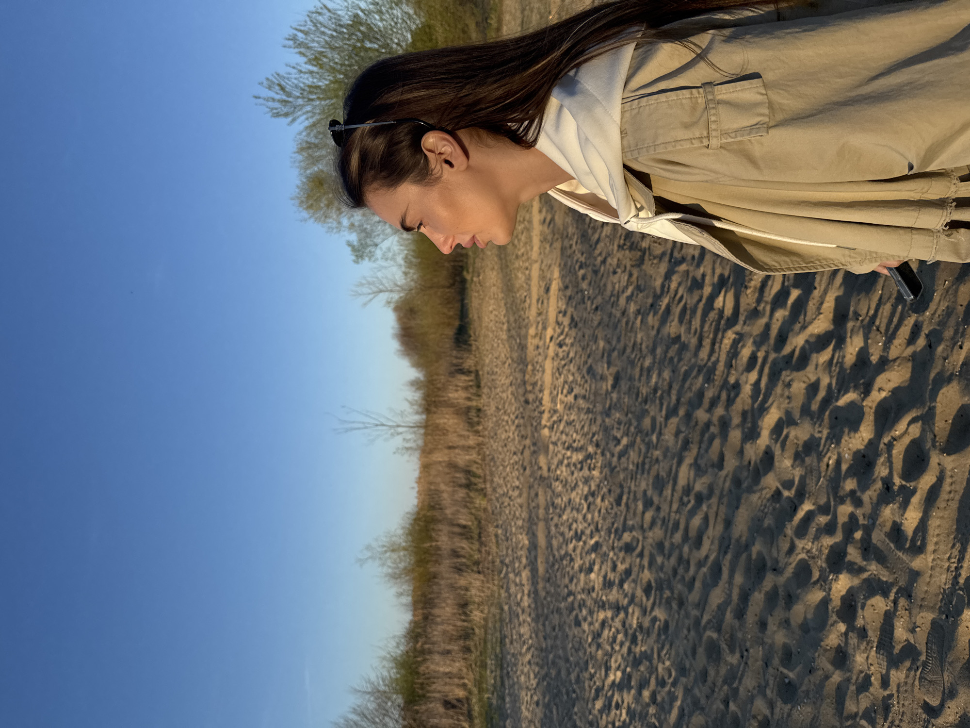 A medium shot of a woman outdoors with a sandy surface and sparse vegetation. The woman has long, dark hair, and wears sunglasses perched on her head. She is wearing a beige jacket over a white hooded sweatshirt. She is looking downwards and her face is visible in profile. The sky is a clear blue. The background includes a sandy surface with patterns of light and shadow, indicating tire tracks. Sparse vegetation, including low-lying plants and small trees, is visible in the middle ground. The location is likely Arciechów, Poland, based on the provided prompt.