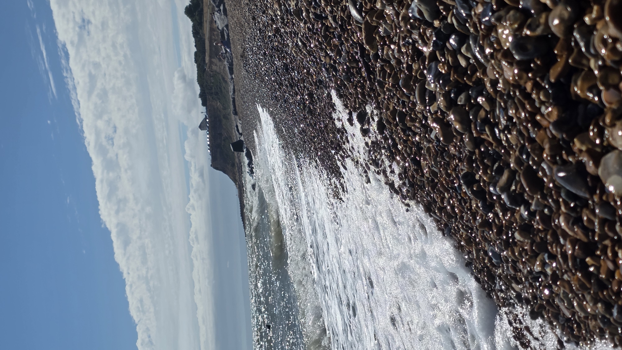 The image depicts a pebble beach with gentle waves lapping the shore. The foreground is dominated by a dense scattering of smooth, rounded pebbles of various shades of brown, grey, and black, many of which are wet and glistening from the sea spray. A line of white foam from the waves marks the edge of the water on the pebble beach.
In the middle ground, the sea stretches out, its surface shimmering under a bright sky. The water appears a deep blue-grey, with whitecaps visible as the waves break. To the left of the frame, the coastline curves away, with a cliff face rising in the background.
On top of the cliff, several structures are visible. One appears to be a building with a distinctive angular roof, possibly a contemporary structure or a former industrial building. A row of parked cars is also present on the cliff top, suggesting a public area or viewpoint. The cliff itself is covered in greenery and rough terrain. The sky above is a mix of bright blue with scattered white clouds, some of which are streaky and elongated, indicating wind at higher altitudes.
In the far background, the horizon is marked by a hazy line where the sea meets the sky. The overall scene is one of a natural coastal environment, with elements of human presence on the clifftop. While the specific location is St Leonards, United Kingdom, no specific landmarks within the city are identifiable from the image alone, beyond it being a coastal area with cliffs. The image captures a sunny day with a moderate sea. There are no discernible people clearly visible in the image, though two figures appear as small shapes on the rocks in the background.