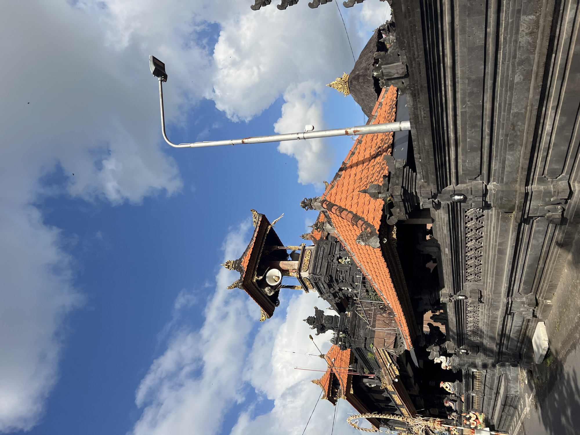 A photograph captures the exterior of a Balinese temple with traditional architecture under a bright blue sky with scattered clouds. The temple features dark stone walls with intricate carvings, topped with orange tiled roofs. A prominent tower with a large speaker is visible, along with decorative golden elements on some of the roof structures. A modern street lamp stands to the right of the temple. In the foreground, a dark stone wall runs along a paved street, with shadows cast by the sun. The scene appears to be in a street setting, likely in North Kuta, Indonesia, given the architectural style. There are no visible people in the image, and the overall impression is of a culturally rich and visually striking religious site.