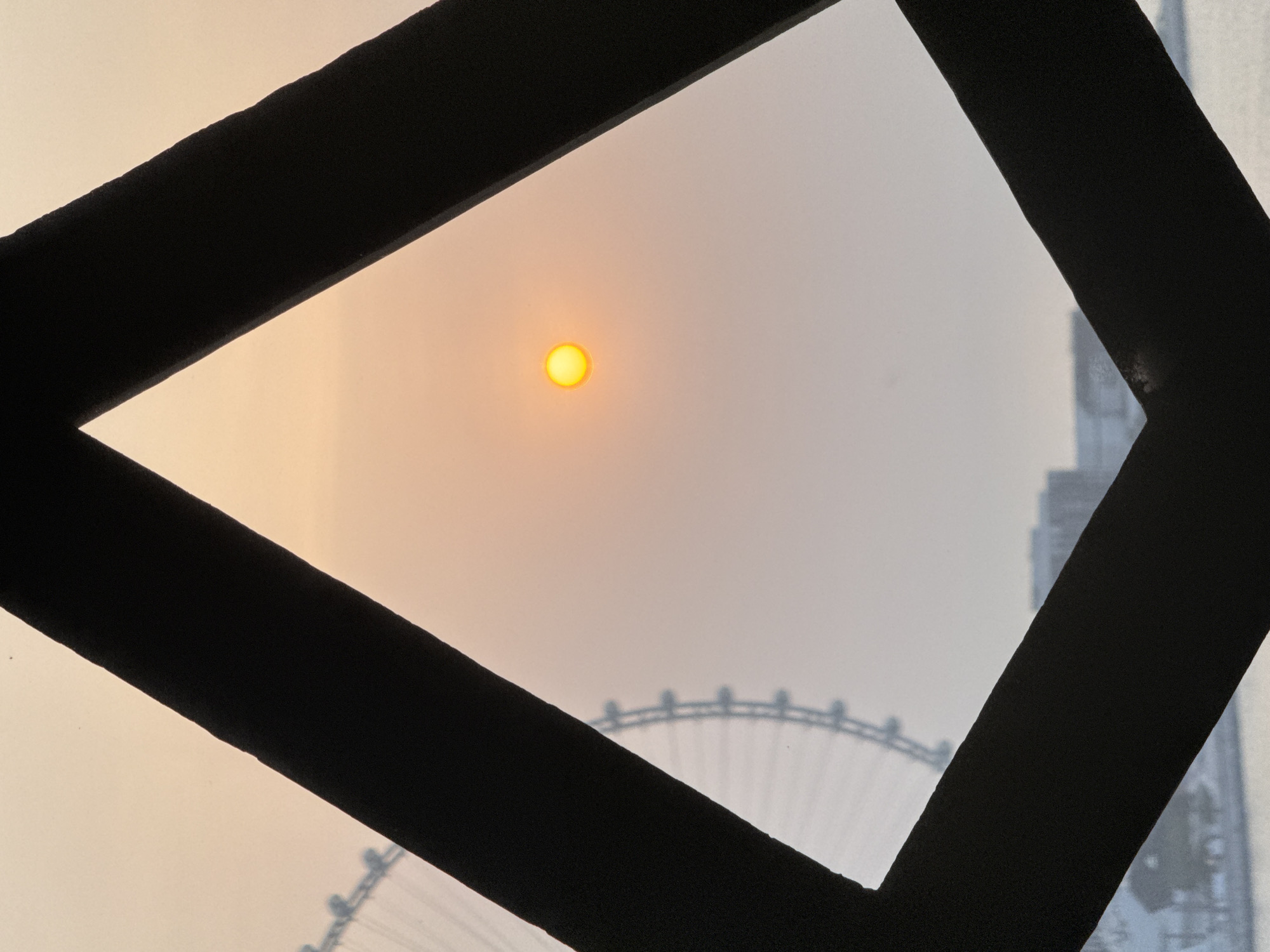 The image shows a hazy sunset viewed through a geometric frame, likely a window or architectural element, in Dubai, United Arab Emirates. The sun, a bright orange disc, is visible in the upper center of the frame, softened by the atmospheric haze. To the left, a portion of a large Ferris wheel, possibly the Ain Dubai, is silhouetted against the sky, its structure partially obscured by the foreground frame. Below the Ferris wheel, a faint outline of Dubai's cityscape is visible, including buildings and what might be water. The framing elements are dark, solid shapes, forming a rough diamond or kite shape with the point facing upwards, effectively isolating the view of the sun and the distant landmarks. The overall impression is one of a filtered, atmospheric perspective on a Dubai sunset.
