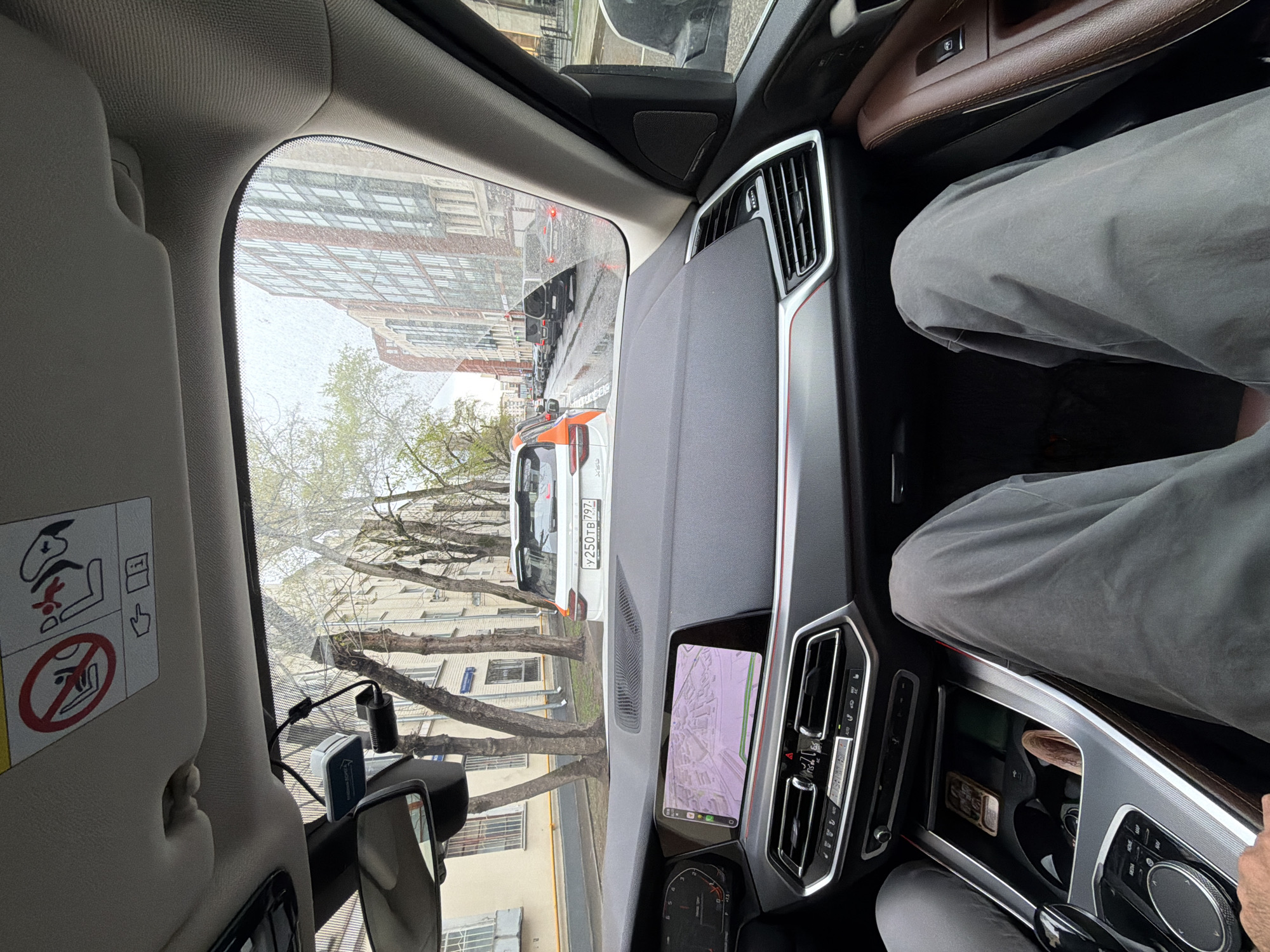 The image shows the interior of a car, a BMW, from the passenger seat perspective, with a view out onto a street in Moscow, Russia. The view outside the car features a street with buildings lining the road and trees with bare branches. A white and orange utility vehicle, an X6, is visible ahead, with a license plate number partially readable as "У 250ТВ 197". Other cars are also visible in the distance.
Inside the car, the dashboard and various controls are visible. On the passenger side, a person wearing grey pants is visible. A navigation screen is on, showing a map. A rear-view mirror is visible. A dashcam or other device is mounted on the windshield. The car's interior is brown and black.