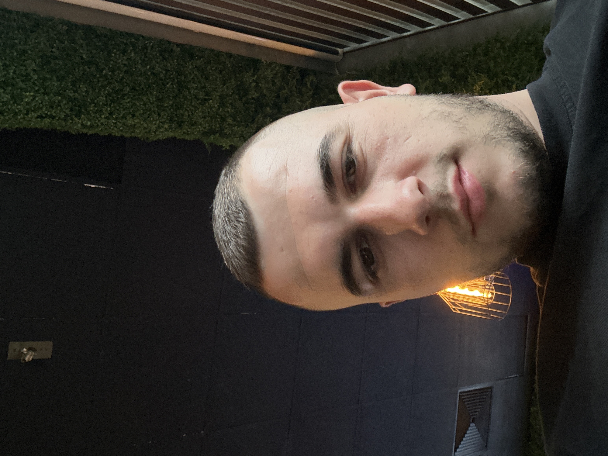 A man with short, dark hair and a beard is looking directly at the camera. He is wearing a dark shirt. The background includes green vegetation, black walls, a light fixture, and a slatted roof. The picture appears to have been taken outdoors in a public space. The location in the city of Plovdiv, Bulgaria is unknown.