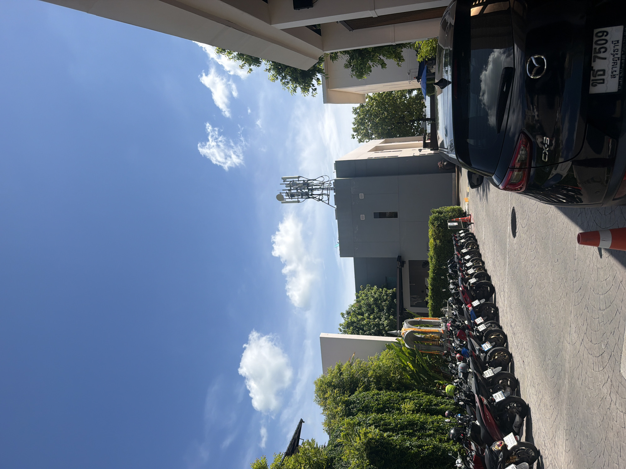 The image depicts a sunlit outdoor scene at Baan Chaweng, Thailand, with a bright blue sky and scattered white clouds. In the foreground, a black Mazda CX-3 is parked, its rear visible. To the left, a long row of motorcycles is parked on a patterned paved surface. Beyond the motorcycles, lush green trees and vegetation line the area. In the mid-ground, a cellular tower is situated on top of a grey building. Further back, another building with a white facade is visible. The overall environment suggests a tropical urban setting with a clear day.