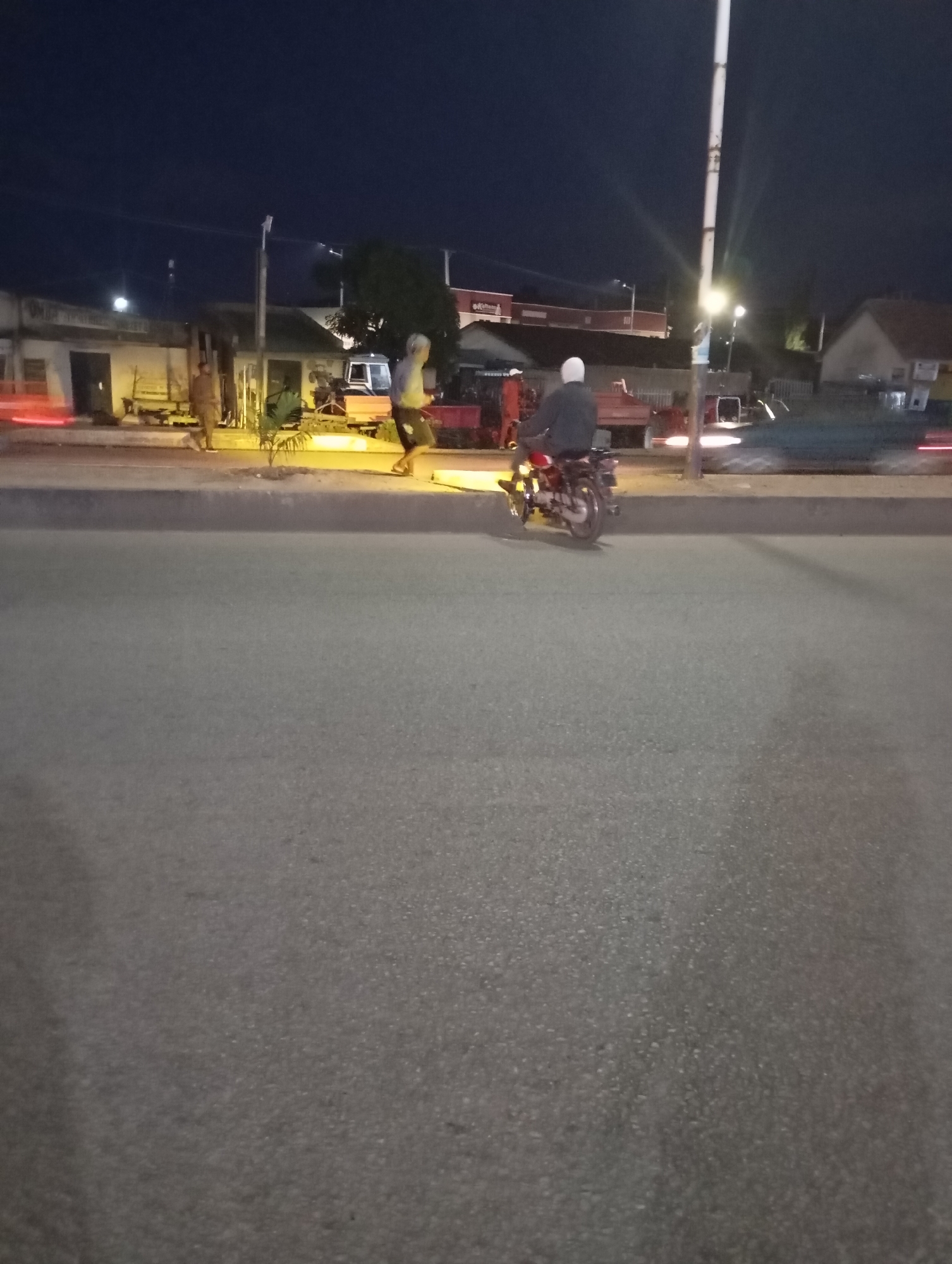 A low-light, eye-level shot captures a street scene at dusk, possibly in Jos, Nigeria, characterized by a dark sky and ambient artificial lighting. A person is seated on a red motorcycle in the foreground, facing away from the camera. The motorcycle is positioned on the right side of the frame, with its rear wheel in focus. In the background, a few figures are visible on a sidewalk in front of buildings. One individual appears to be running or in motion, while another stands. Beyond them, a truck and other vehicles are present, their forms blurred by the low light and motion. The overall impression is of a quiet moment on a street as night falls, with subtle hints of activity.