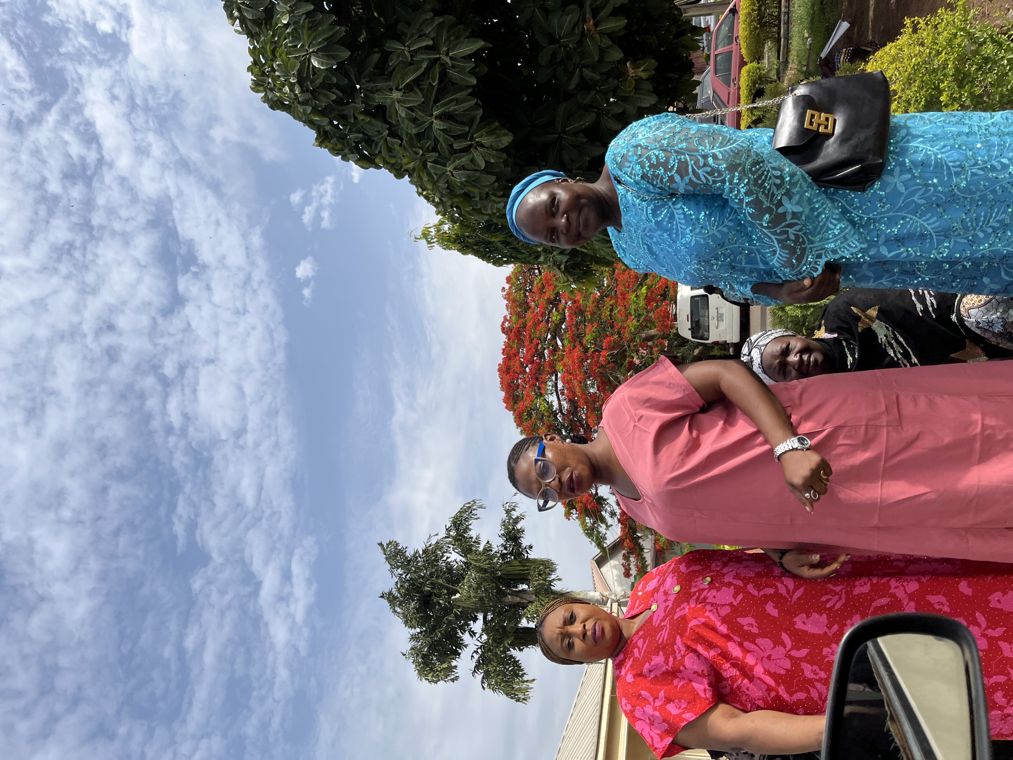 The image shows four women standing outdoors on what appears to be a sunny day. The woman at the top is wearing a blue lace dress and has a black purse over her shoulder. The woman in the middle is wearing a pink dress and eyeglasses. The woman at the bottom is wearing a red dress with a pink floral pattern. There is a fourth woman behind the woman in the blue dress. The environment includes trees, a sky with clouds, what appears to be a vehicle, and part of a building. Based on the information in the original prompt, the location is Jos, Nigeria. No specific actions or interactions are evident other than them standing together.