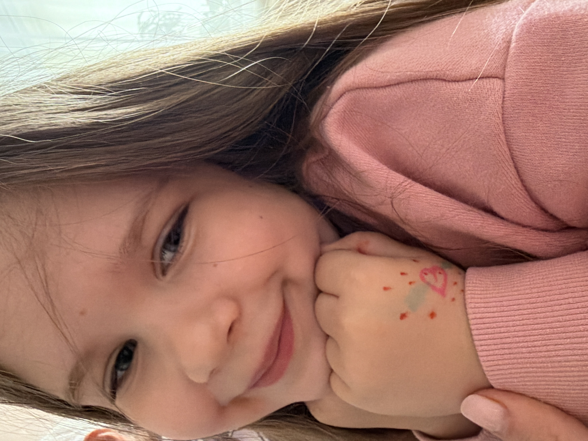 In Valozhyn, Belarus, a young girl with long, light brown hair is captured in a close-up, portrait-style photograph. She is leaning on her hands, which are positioned at her chin, and she is smiling gently, with her eyes looking slightly off-camera. Her left hand has a drawing on it, appearing to be a pink heart with some red dots and a light blue shape surrounding it. She is wearing a pink sweatshirt with ribbed cuffs. The lighting suggests an indoor setting, possibly near a window due to the soft, natural light illuminating her face and hair. The background is out of focus, with hints of a pale, possibly white or light-colored wall or curtain.