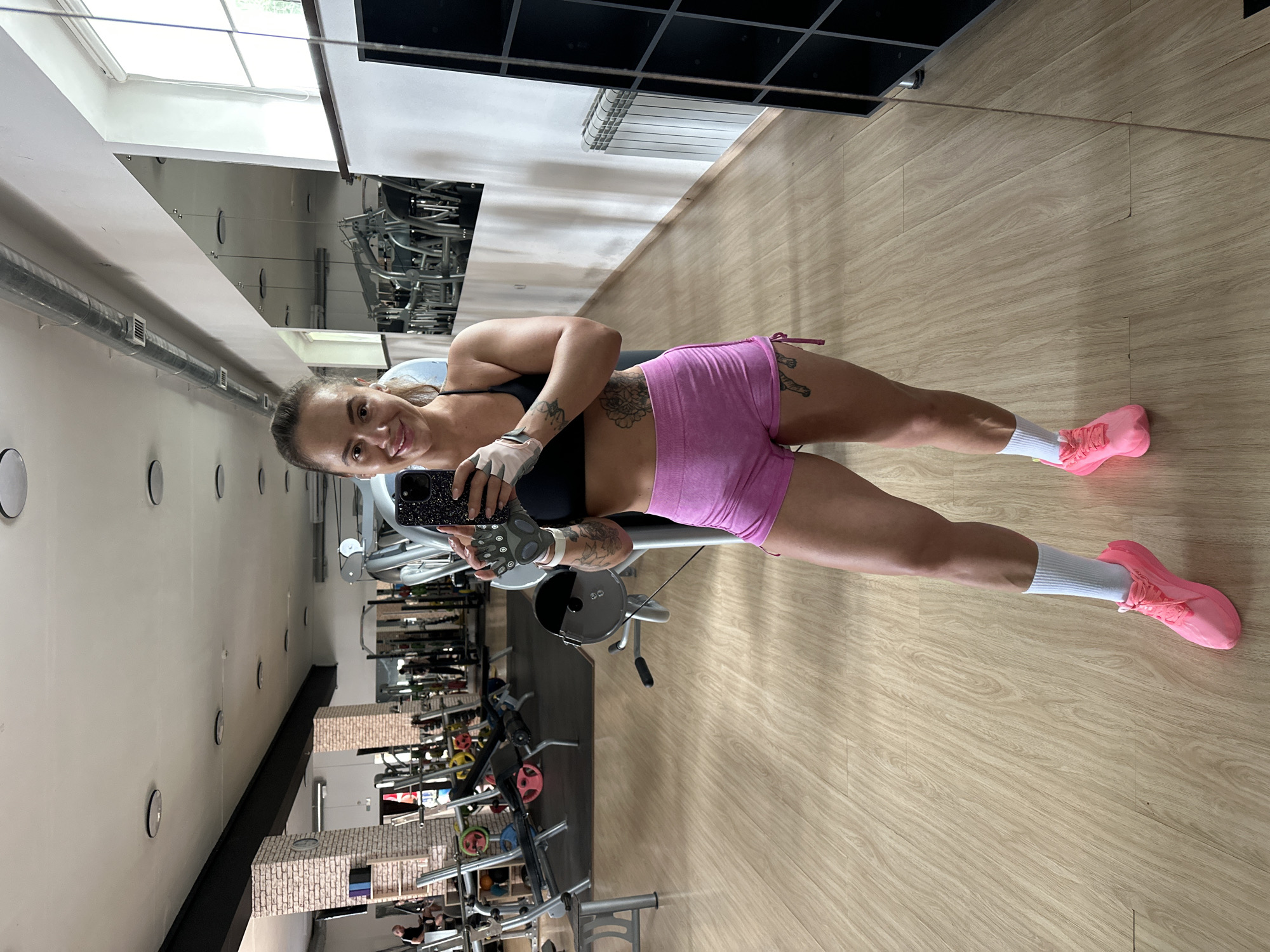 A full-body selfie is taken by a woman in a gym. She is wearing a black sports bra, pink shorts, white socks, and pink sneakers. She has on workout gloves and is holding a smartphone in her right hand. She has tattoos on her left arm and leg. The gym has wooden floors, gym equipment, and mirrors. The location is Yuzhnouralsk, Russia.
