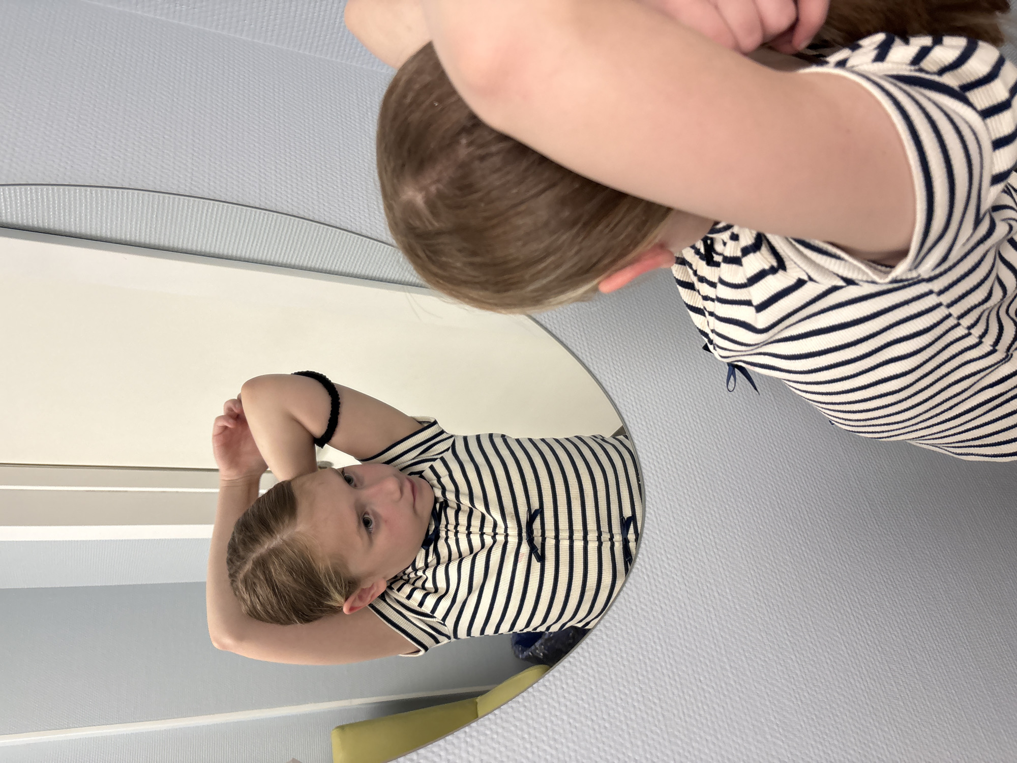 A young girl is shown looking at her reflection in a large, round mirror. The image captures both the real girl, seen from behind, and her mirrored image.
The real girl, positioned in the upper right of the frame, has light brown hair parted down the middle. Her arms are bent, with her hands resting near her head, suggesting she might be adjusting her hair or simply resting. She is wearing a short-sleeved, horizontally striped shirt or dress in white with dark blue or black stripes.
Her reflection in the mirror, which occupies the left side of the frame, shows her face in profile, looking towards her left. She has light-colored eyes and a neutral, contemplative expression. The striped garment she wears is visible, adorned with small dark blue bows on the front. A black hair tie or bracelet is on her left wrist.
The setting is indoors, featuring light blue/grey textured walls. A lighter surface, possibly a counter or floor, runs below the mirror. In the mirror's reflection, a glimpse of a yellow object, likely furniture, is visible at the bottom. The girl appears to be engaged in self-observation or grooming. No text is visible in the image. The location context provided is Veliky Novgorod, Russia.