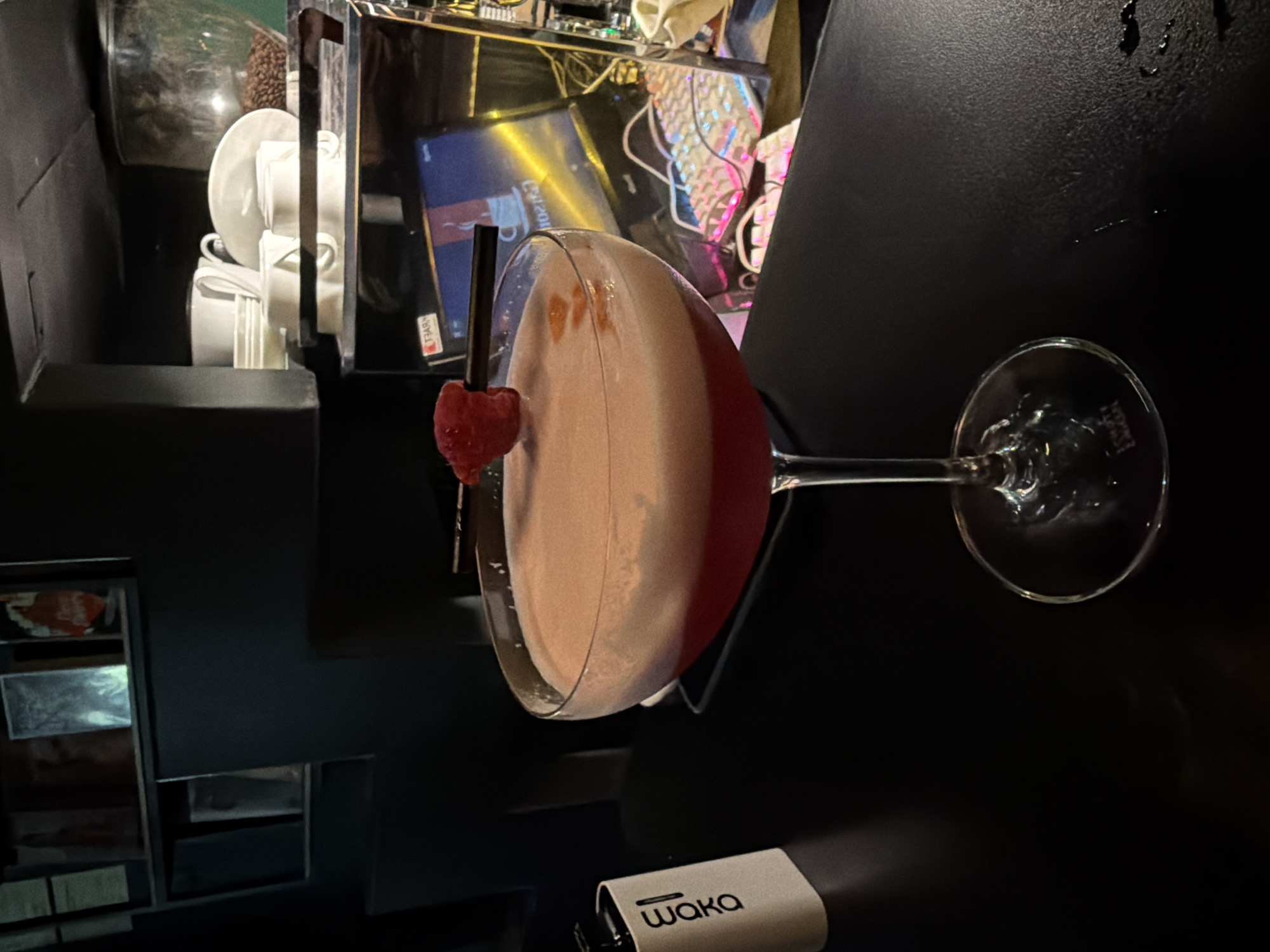 In Budva, Montenegro, a cocktail glass containing a frothy, pink beverage with a raspberry garnish sits on a dark, reflective surface, likely a bar counter. A small white object with the text "waka" is visible to the left of the glass. In the background, shelves hold various items, including what appear to be books or records. Further back, a coffee machine with a glass hopper filled with beans is present, along with stacked white cups and saucers. A computer monitor displaying an image of a coffee cup and a keyboard with colorful backlighting are also visible in the background, suggesting a contemporary bar or cafe setting. The lighting is dim, creating a moody atmosphere.