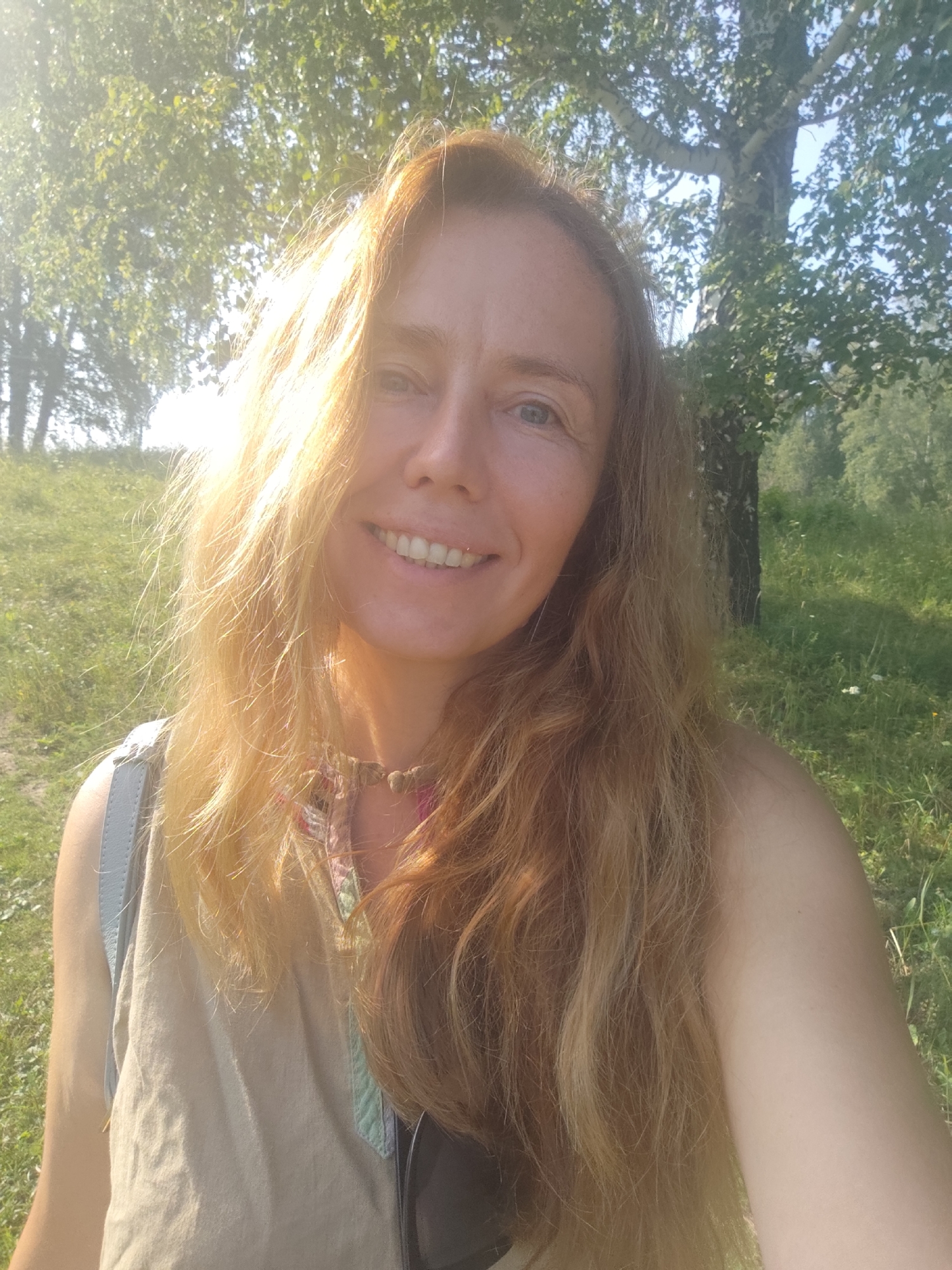 A woman with blonde hair smiles at the camera in a selfie taken outdoors. She is wearing a light-colored, sleeveless top and a necklace. A light blue strap, likely from a backpack, is visible over her left shoulder. The background consists of a grassy slope, trees, and sunlight filtering through the leaves. The image appears to be taken in a park or natural area, potentially in Oktyabrskiy, Russia, given the context.