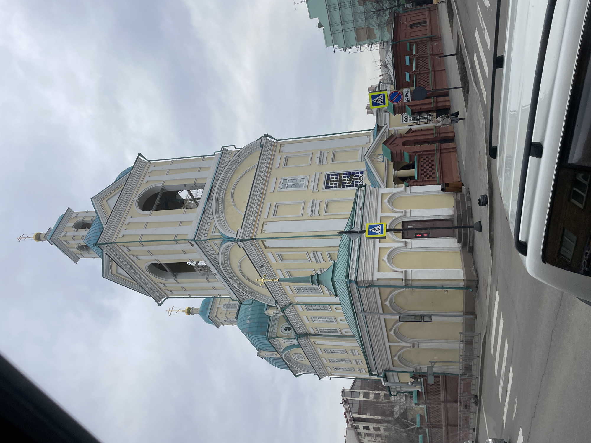 The image, correctly oriented, depicts a street scene dominated by a large, ornate church on the left side. The church features light yellow walls with white architectural details and multiple teal-colored domes, each topped with a golden Orthodox cross. A prominent, multi-tiered bell tower, also in yellow and white, rises above the main structure, culminating in a dome and cross. The architecture includes classical elements such as pilasters, arched windows, and intricate cornices.
In the foreground and to the right, a paved road with a white "zebra" pedestrian crossing is visible. A white vehicle, likely a van or SUV, is partially seen on the far right, suggesting the photo was taken from a car or roadside. A sidewalk runs along the front of the church. Across the street, to the right of the church, stands an older brick building with dark windows, and scaffolding on a building behind it hints at construction or renovation.
One pedestrian, dressed in a light-colored coat, is walking on the sidewalk near the brick building. Several street signs are visible: two blue square signs with white triangular pedestrian symbols (pedestrian crossing ahead) and a circular red sign with a white horizontal bar (no entry). A black trash bin is located on the sidewalk near the church entrance.
The scene takes place on an overcast day, indicated by the uniformly grey and cloudy sky. The lighting suggests it is daytime. The architectural style of the church and the design of the street signs are consistent with a Russian urban setting, specifically Krasnoyarsk as per the context. No clear legible text is visible other than the symbols on the street signs; red double arrows appear on a church door. The image captures an everyday street scene with a focus on the historic church.