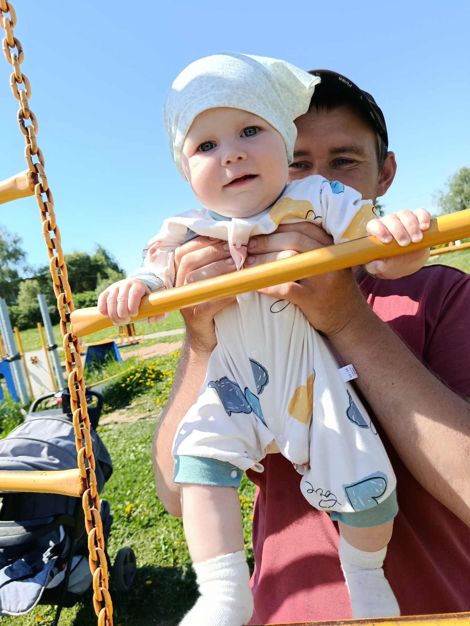 A baby in a white patterned onesie and a light blue hat is being held up by an adult male at a playground on a sunny day in Фоки, Russia. The baby is holding onto a yellow horizontal bar attached to a yellow chain. The adult is wearing a maroon shirt and a black baseball cap. A gray stroller is partially visible in the background. The playground is surrounded by grass and trees under a clear blue sky. Other playground equipment is visible in the background.
