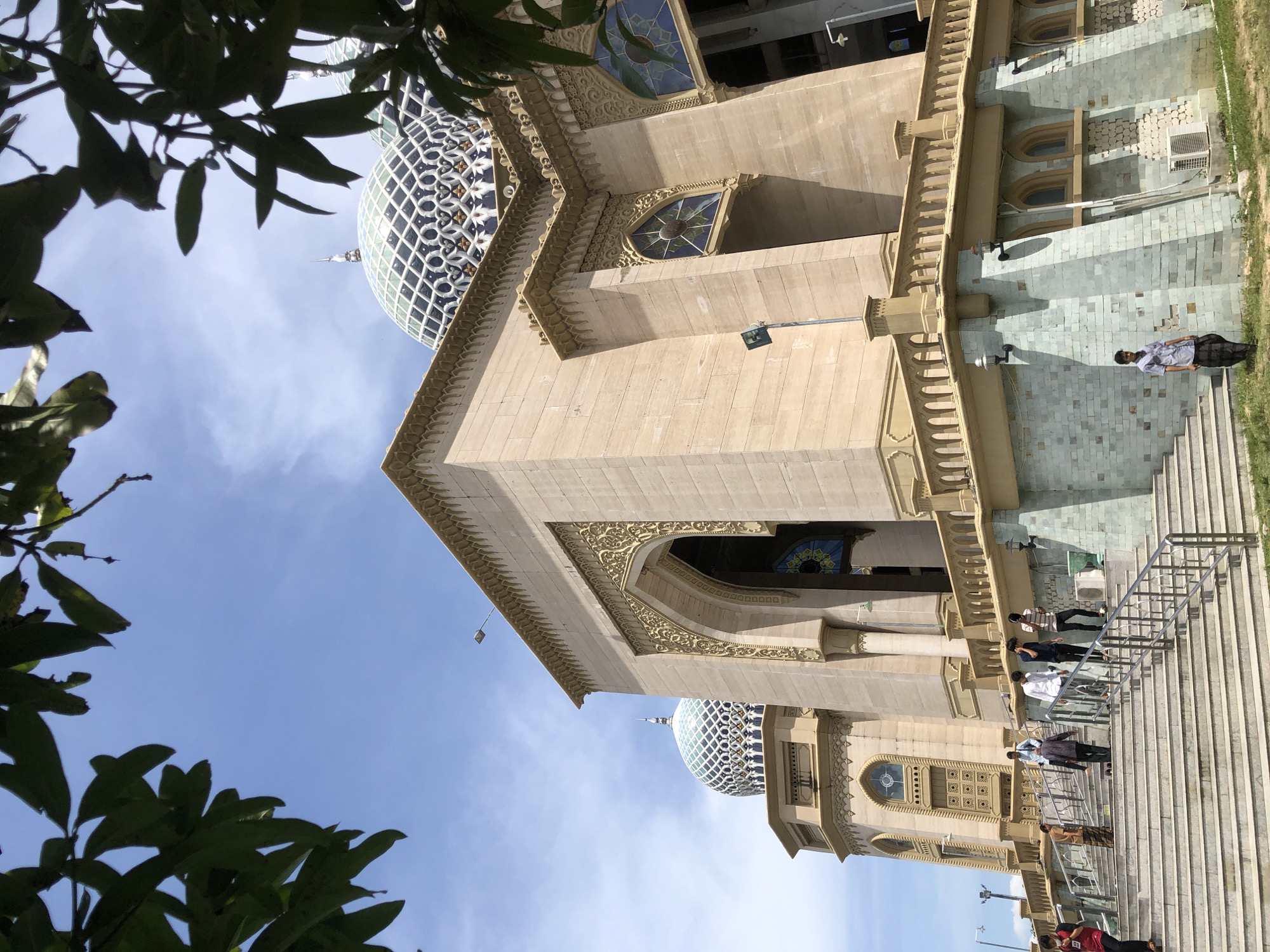 The image depicts the Baiturrahman Grand Mosque in Lhokseumawe, Indonesia. The mosque is a beige-colored building with ornate carvings and a blue and white dome. A set of stairs with a metal railing leads to the mosque's entrance, where several people are standing. The sky is blue with some clouds. A leafy tree partially obscures the view from the left side of the frame.