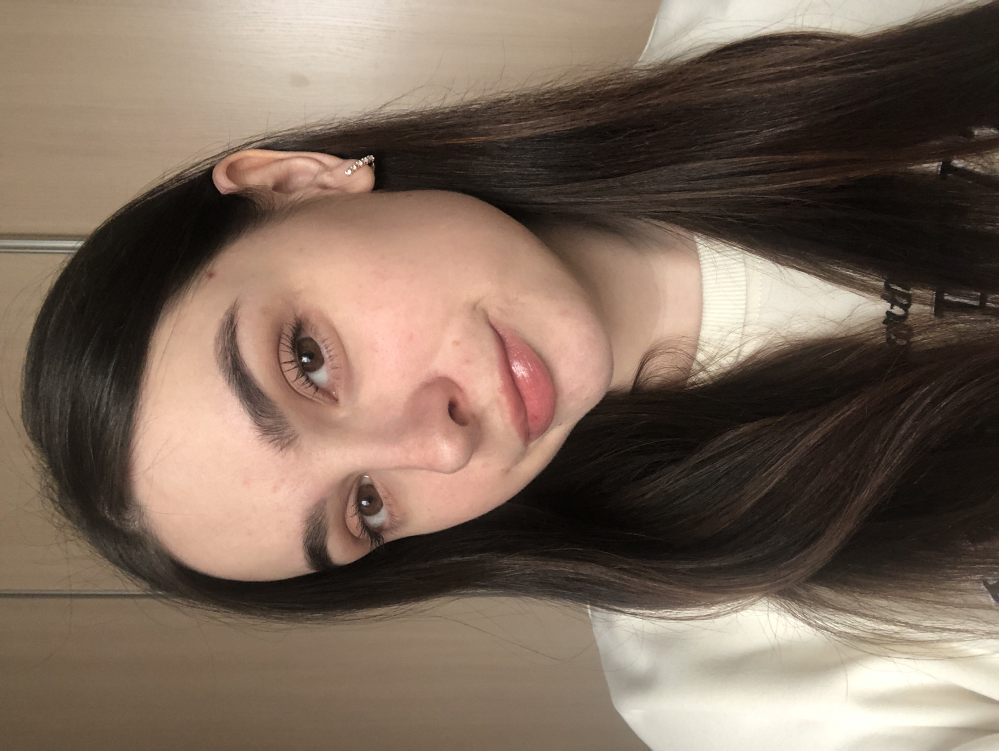A close-up portrait of a young woman with long, dark brown hair. She is wearing a white sweater. Her skin is fair with rosy cheeks. Her eyes are brown and her eyebrows are thick. She has a slight smile. A small, jeweled earring is visible in her left ear. The background is a light-colored wall. The image does not contain any information about the city of Noviy, Russia.