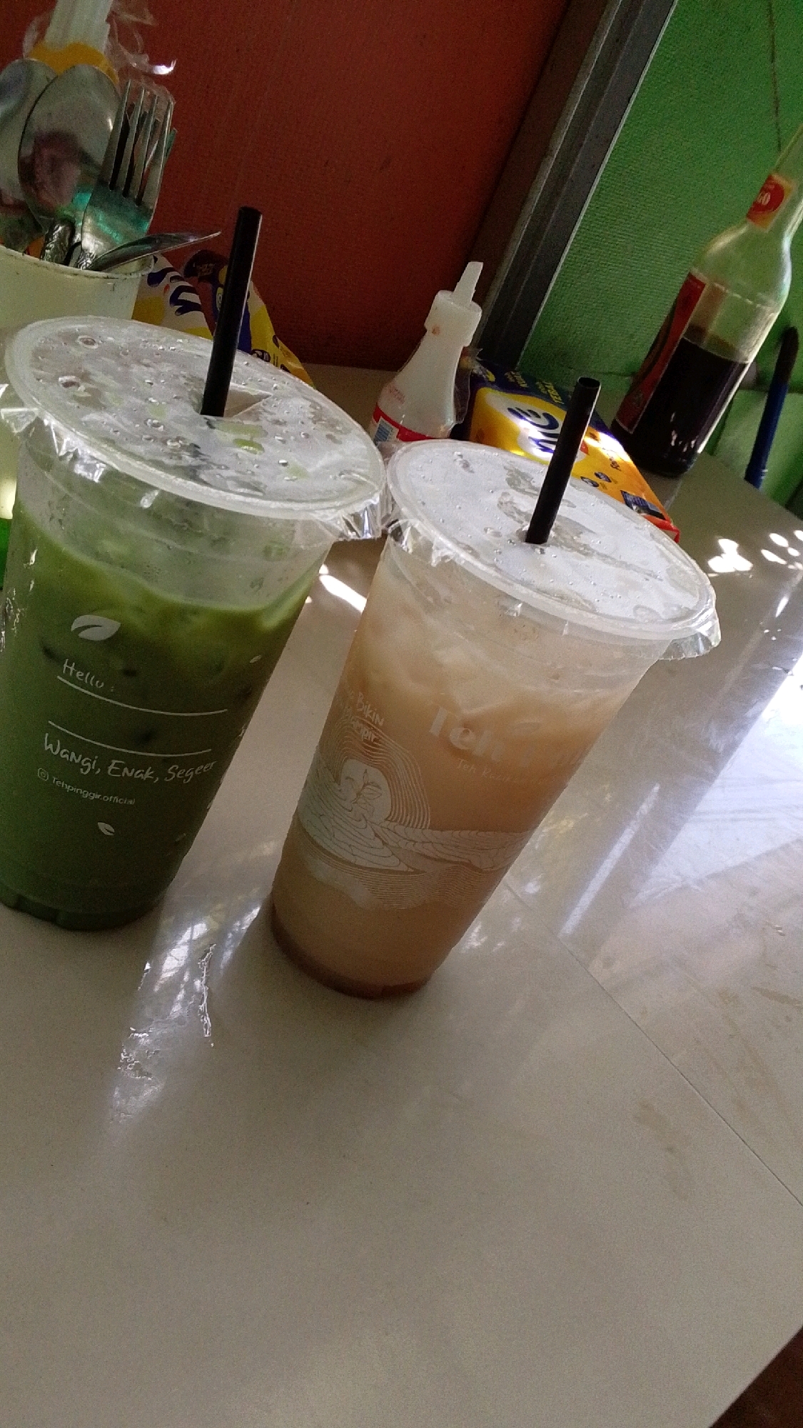 The image depicts two plastic cups with clear lids and black straws, each filled with a beverage. The cup on the left contains a green liquid, likely iced matcha, with text that reads "Hello: Wangi, Enak, Seger @Tehpinggir.official". The cup on the right contains a lighter colored, foamy beverage, possibly iced tea or a milk-based drink, with some text on the cup that is partially obscured but includes "Bikin" and "Teh". In the background, to the left, is a white container filled with cutlery, including spoons and forks. Behind the cups, there are several items: a white squeeze bottle with a red cap, a yellow and blue packet, and a glass bottle with dark liquid. The setting appears to be an indoor environment with a tiled surface reflecting light, suggesting it might be a table or countertop. The overall lighting suggests daytime, with shadows cast on the surface. The background walls are painted in shades of reddish-brown and green. This image does not provide enough information to determine the specific location in Cintaratu, Indonesia, or any details about people, actions, or interactions.