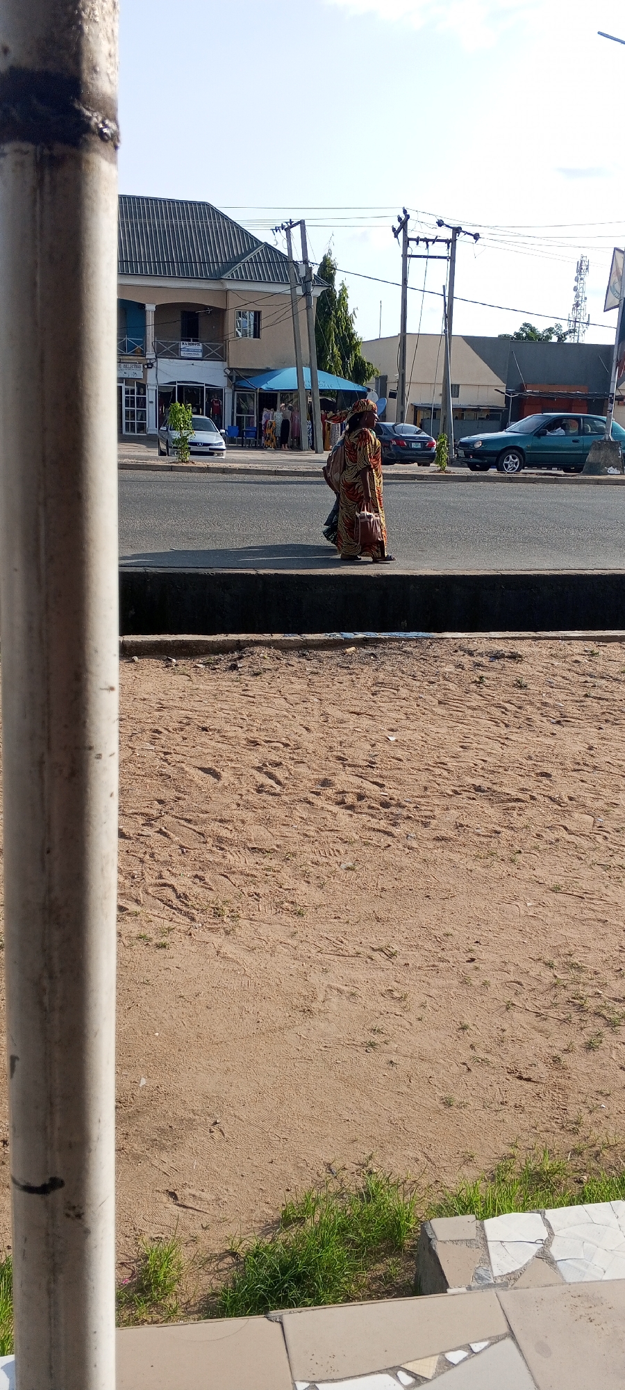 The image shows a street scene in Jalingo, Nigeria. In the foreground, a portion of a light-colored, textured pole is visible on the left side, obscuring a portion of the scene. The middle ground features a paved road with a raised concrete curb. A person, likely a woman, is walking along the curb, wearing a colorful, patterned robe and carrying a large bag. Behind her, several cars are parked and moving on the road, including a green sedan and a darker sedan. The background consists of buildings lining the street, with shops visible at ground level. One shop has a blue awning, and displays clothing. Overhead, power lines and utility poles are present, and a cellular tower is visible in the distance. The ground on the right side of the curb appears to be unpaved and sandy with sparse patches of grass. The sky is clear with some light clouds.