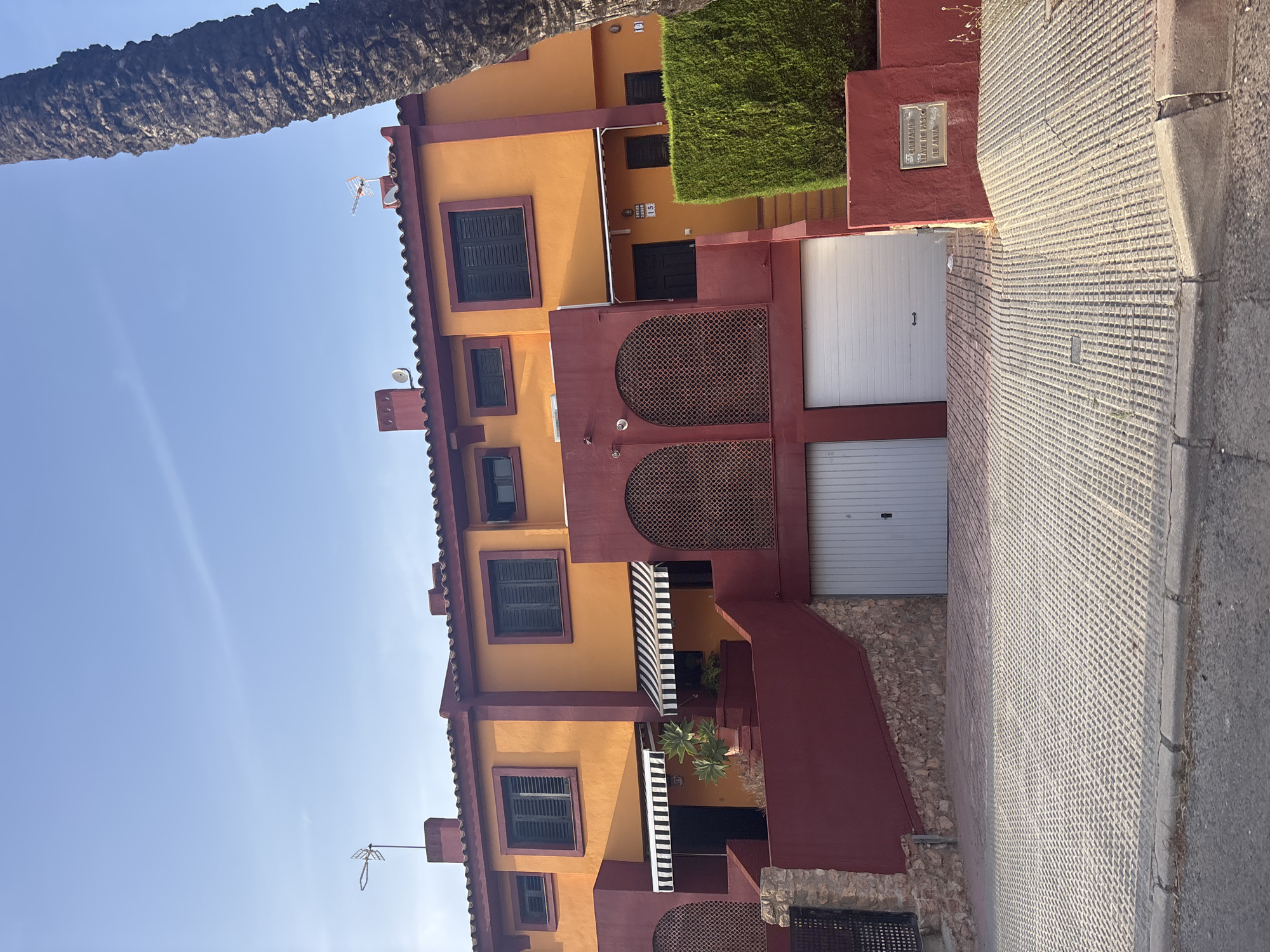 The image depicts a row of modern townhouses painted in bright orange with dark red accents. Each unit features dark-shuttered windows, some with black and white striped awnings above the entrances. The houses are built on a slight incline, with driveways leading to garages at the lower level. A thick, textured palm tree trunk partially obscures the right side of the frame. The ground in front of the houses is paved with a grid-like pattern of concrete blocks, and a paved sidewalk separates it from a street. A manicured green hedge is visible to the right of the entrance of one of the houses, next to a red wall with a bronze plaque. The sky is a clear, bright blue. The architecture suggests a Mediterranean style, common in coastal areas of Spain. Given the distinct architectural style and the context of Orihuela, Spain, this scene is likely situated in a residential area within or near the city, possibly in a development designed to maximize space and incorporate private garages.