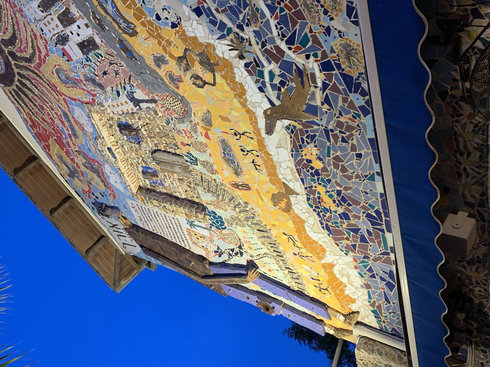 This image shows a mosaic artwork on the exterior of a building in Tel-Aviv, Israel. The artwork is composed of small, irregular pieces of tile in various colors, forming a complex and detailed mural. The colors used in the mosaic include blue, yellow, red, and white. The mural appears to depict scenes of nature, including birds, fish, and other animals. There are also abstract patterns and geometric shapes incorporated into the design. The artwork is mounted on a building and protected by an awning.
