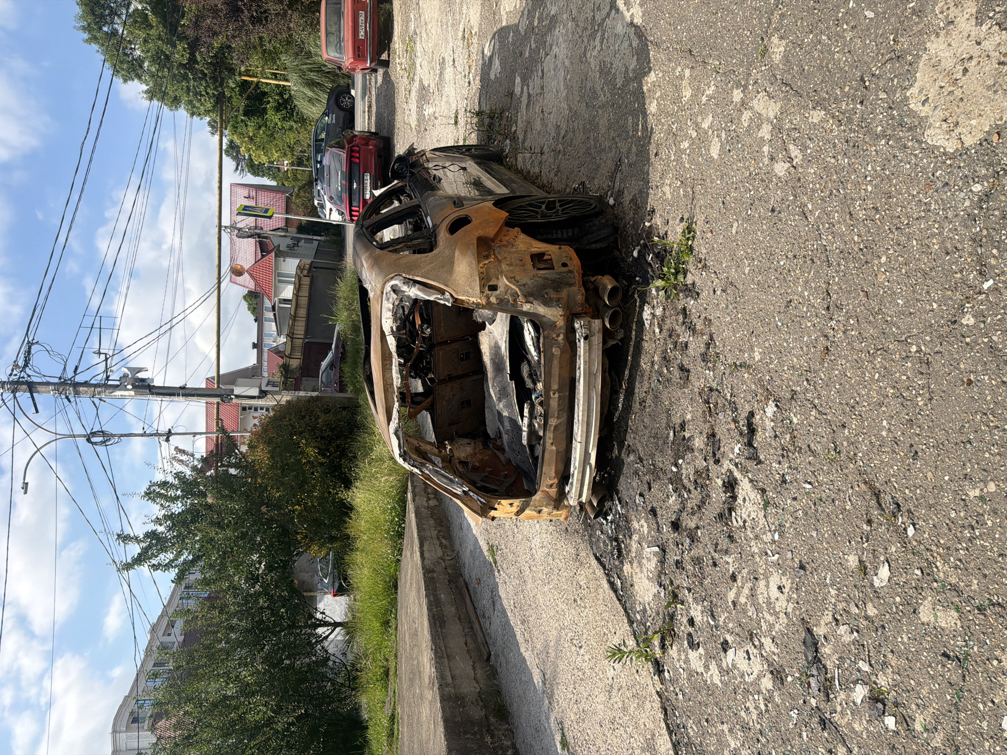 The image shows a burnt-out car on a cracked asphalt road in Sochi, Russia. The car is heavily damaged, with its interior exposed and charred. It appears to be a modern vehicle, possibly a sports car, with dual exhaust pipes visible. The car is lying on its side, suggesting it may have been involved in a serious accident or fire.
The setting is outdoors, with a blue sky and fluffy white clouds visible in the background. There are trees and buildings in the distance, indicating an urban or suburban environment. Power lines and a street light are also present, suggesting it's a public road.
Other cars are parked nearby, including a red sedan and a red sports car, which appear to be intact. The presence of these cars might suggest that the burnt-out vehicle was part of a group or parked among others. The overall scene is desolate and evokes a sense of destruction and loss. There is no visible text in the image. The time of day appears to be daytime, with bright sunlight casting shadows. The weather appears to be clear and sunny.