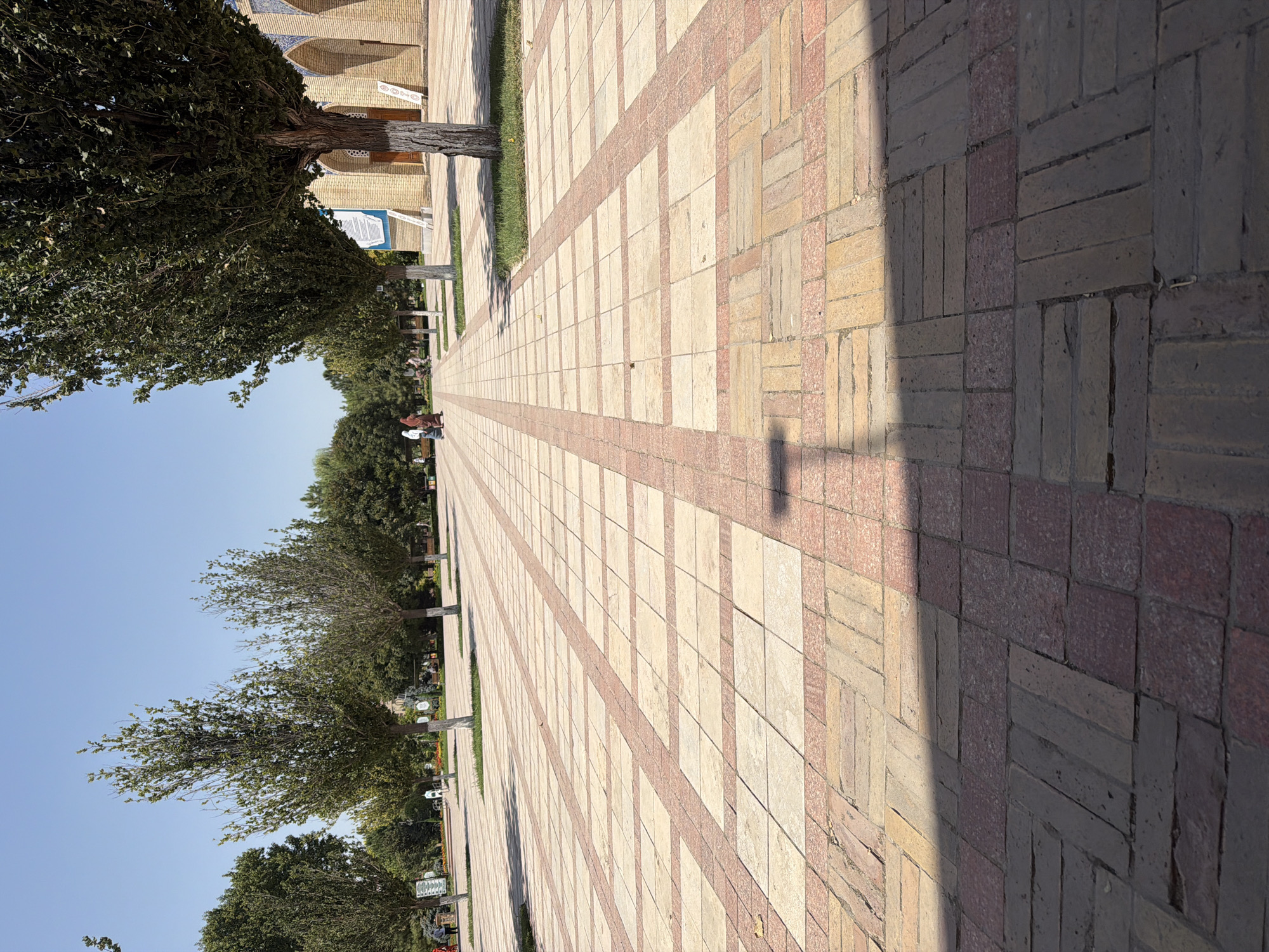 A low-angle, ground-level view of a wide, paved walkway in Zangiata, Uzbekistan. The walkway is constructed with a pattern of alternating light and reddish-brown rectangular tiles. Trees with white-painted bases line the left side of the walkway, interspersed with patches of grass. In the background, to the right, is a building with light yellow brick walls and arched doorways, possibly a mosque or historical site. Several people, dressed in modest clothing, are walking in the distance on the walkway. The sky above is clear and blue, indicating a sunny day. The foreground is dominated by the intricate brickwork of the pathway, with a shadow cast from an unseen object.